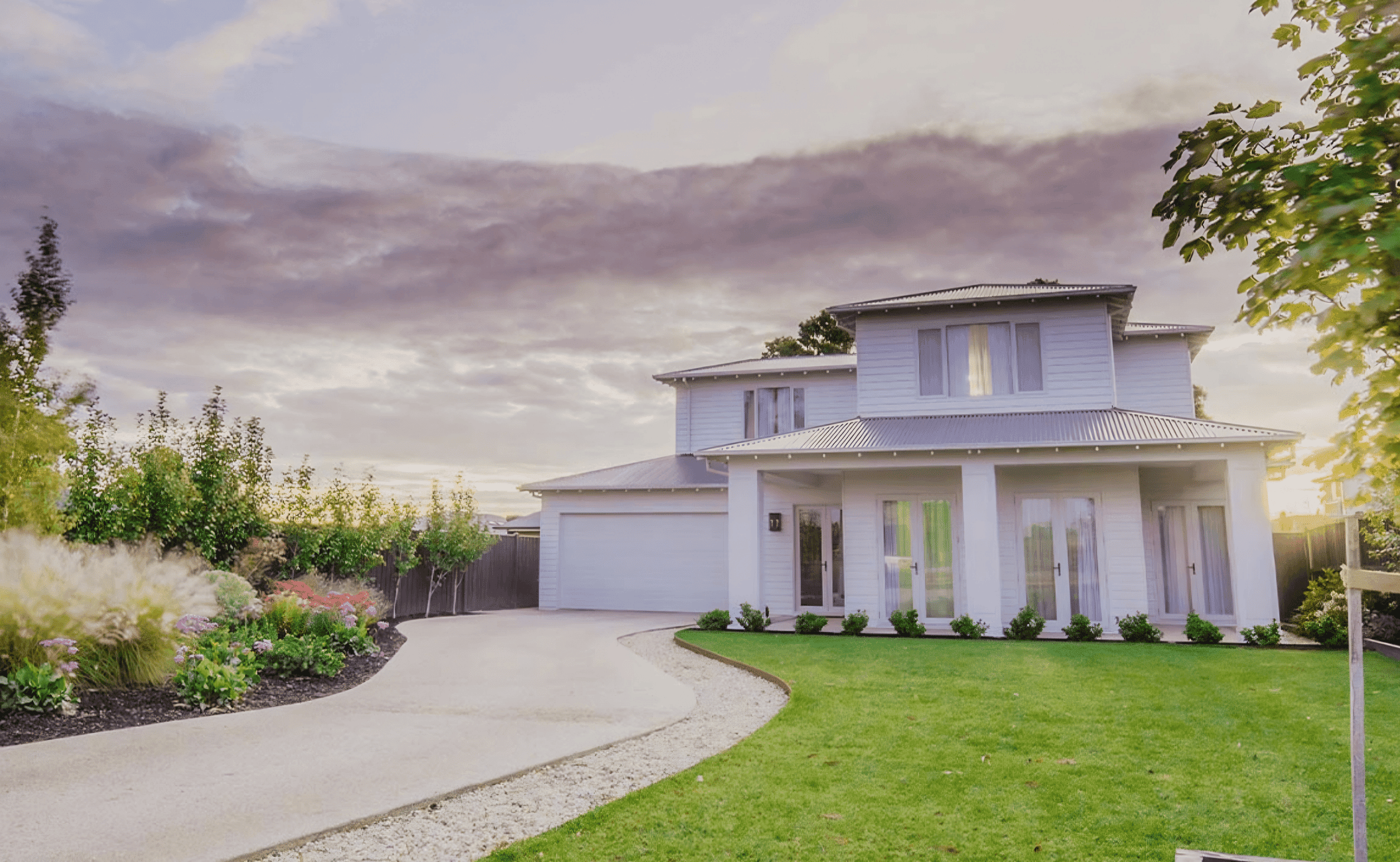 A white two-story house with a front yard featuring a curved gravel driveway, green lawn, and flower bed, under a cloudy sky during sunset.