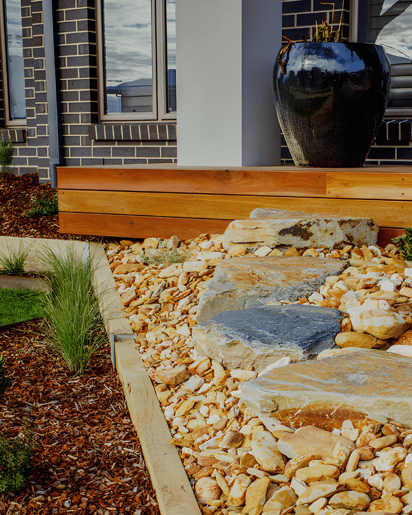 A landscaped garden with large rocks, gravel, and wood border, adjacent to a house with brick walls and window, and a black ceramic pot on a wooden platform.