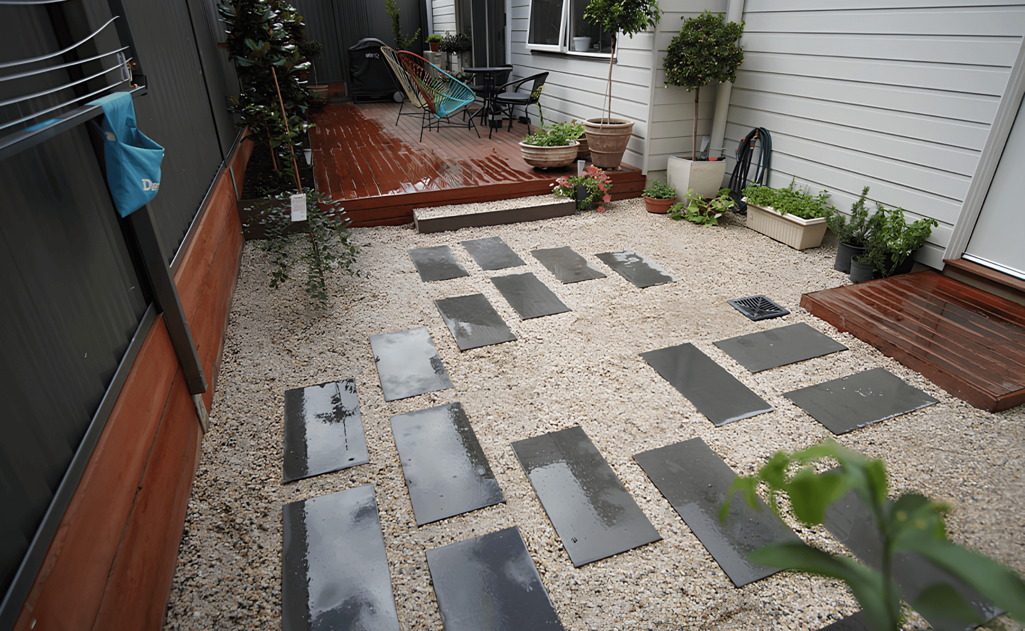 Backyard patio with wet pink wooden deck, potted plants, outdoor furniture, and slate stepping stones on gravel ground.
