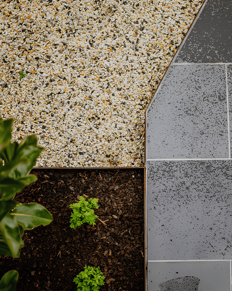 A landscaped outdoor space with gravel, soil, plants, and tiled pavement, separated by a metal edge.