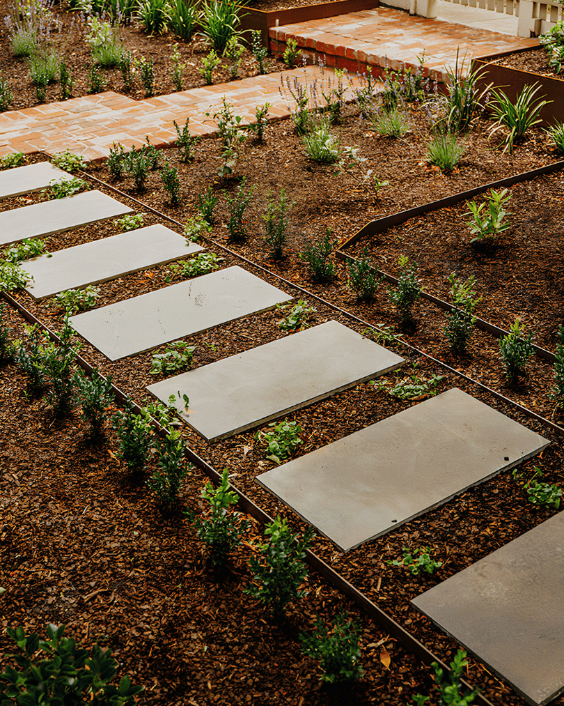 A garden with a brick patio, stepping stones, and various green plants and shrubs.