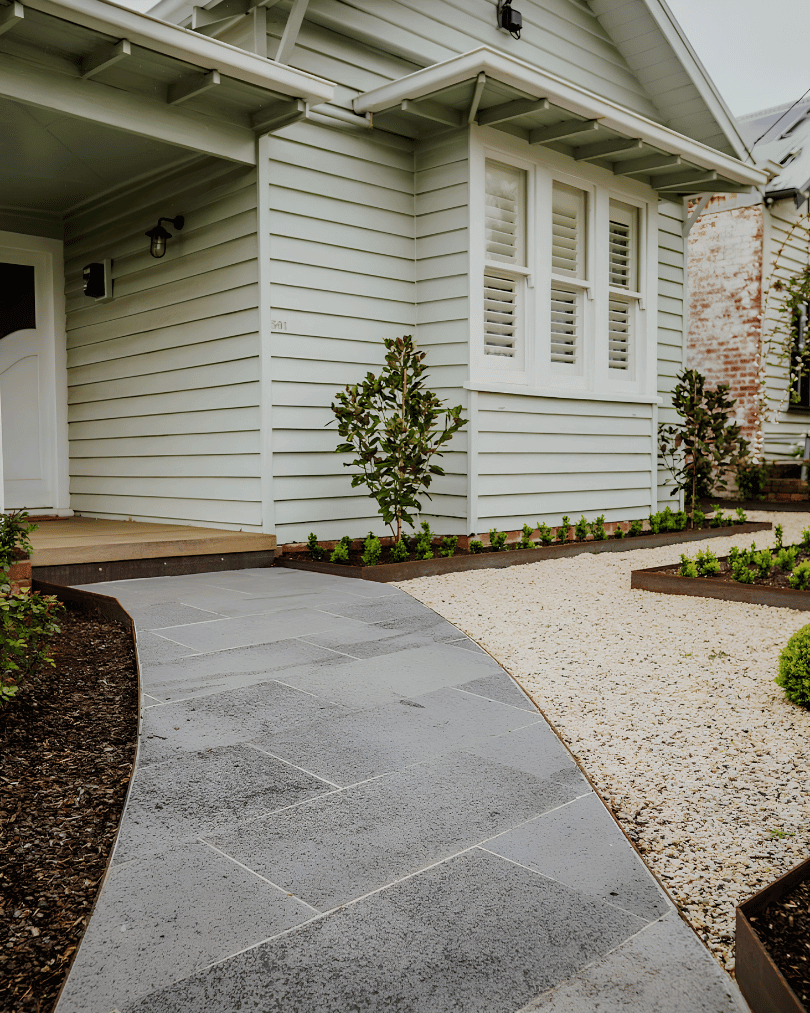 A house with white siding and a window with white shutters, a new gray stone walkway leading to the front door, and a landscaped yard with small plants and shrubs.
