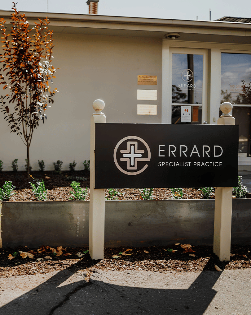 Sign for Errard Specialist Practice in front of a building with a window and a small garden bed with plants and a tree.