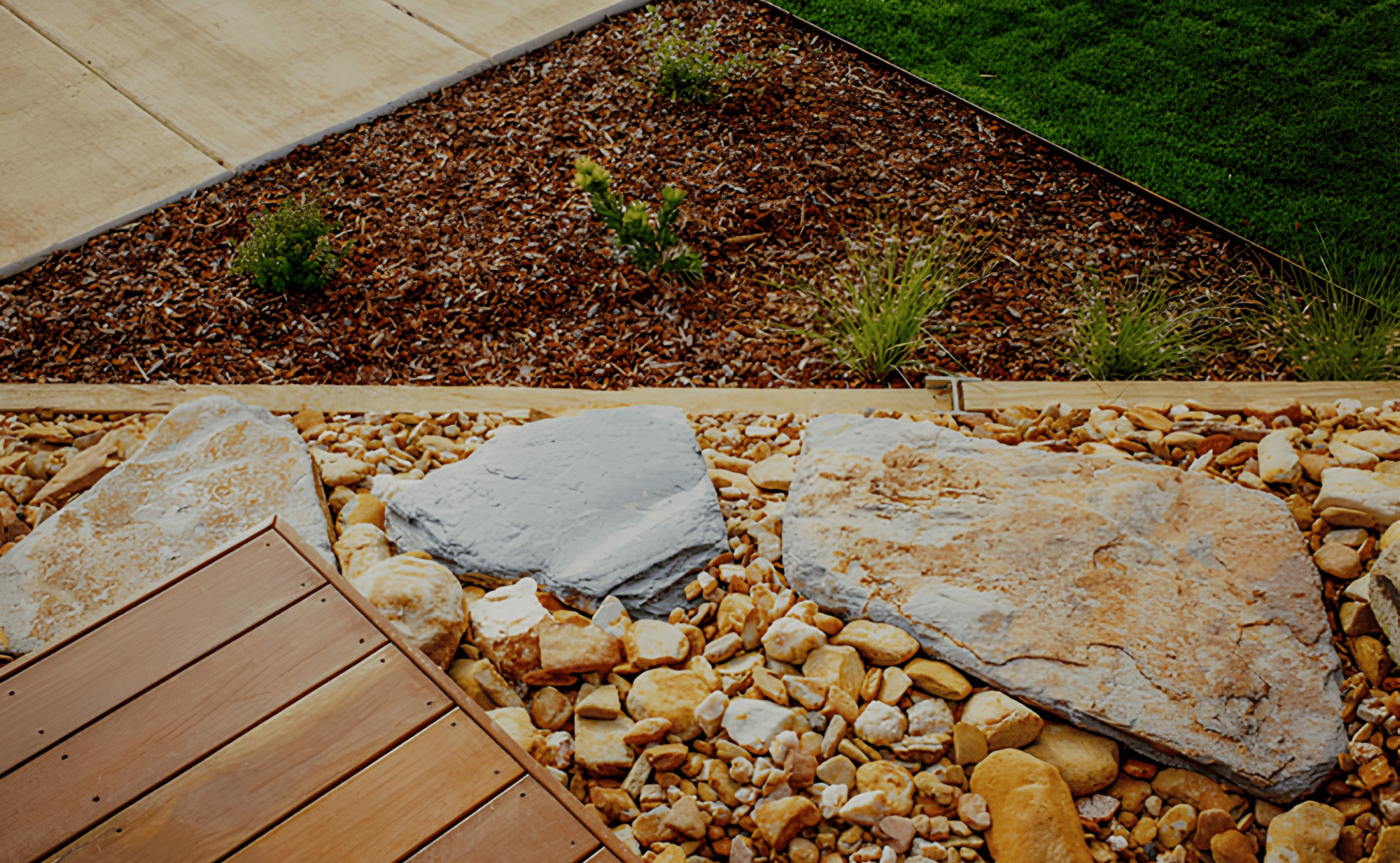 A landscaped garden with a section of brown gravel, small green plants, a wooden border, a small patch of green grass, and a tiled walkway.
