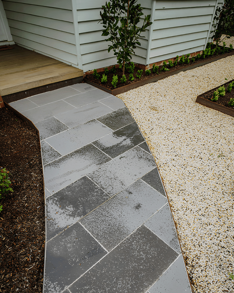 Wet gray stone walkway next to a garden with small plants and a white house wall.