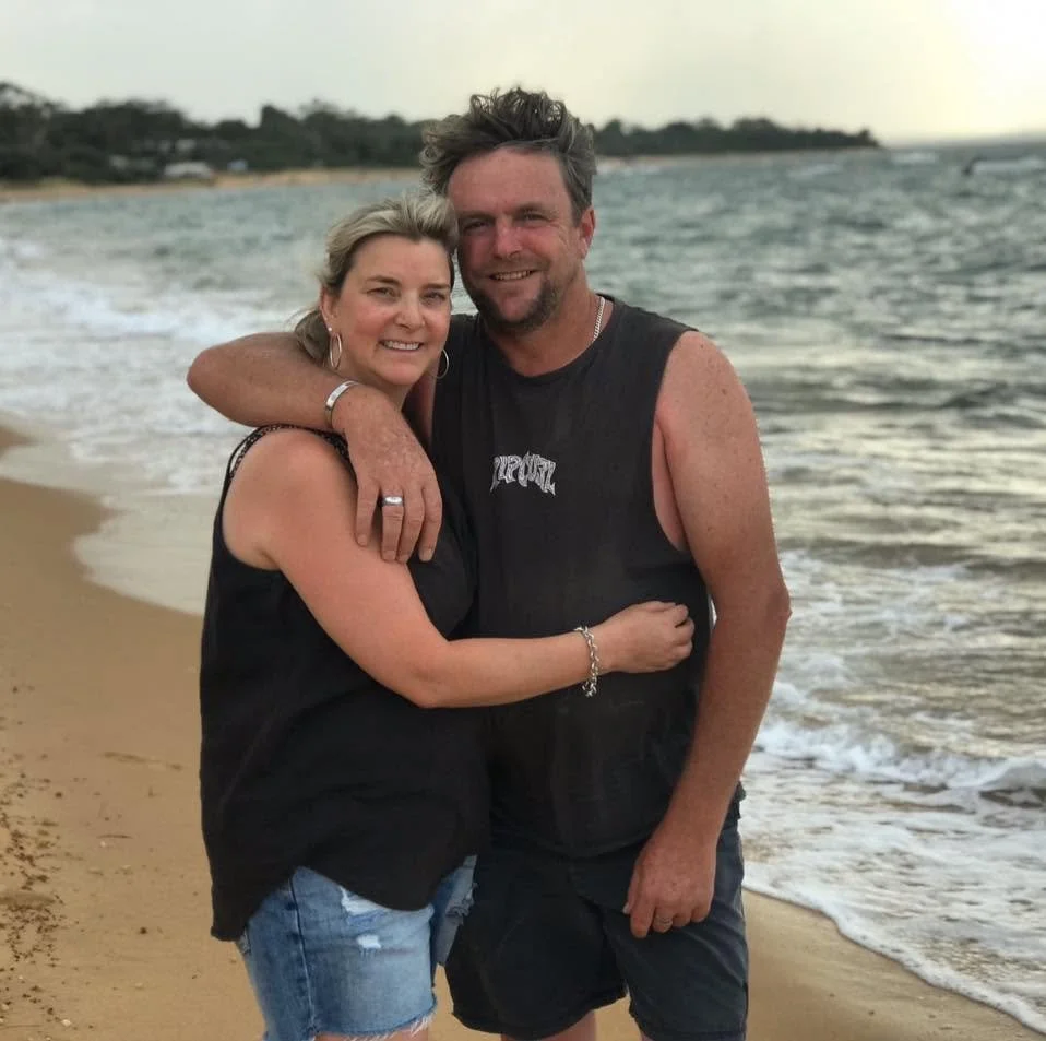 A smiling couple hugging on a beach during sunset, with water and distant land in the background.