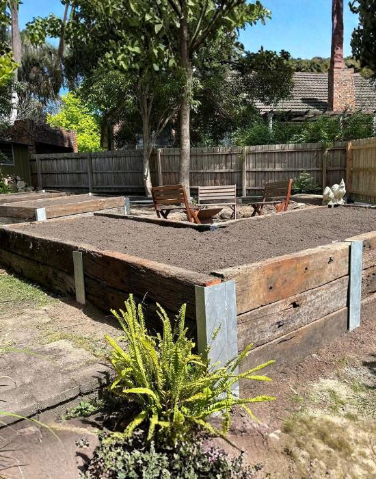 Raised garden bed with freshly tilled soil, surrounded by a wooden border, with three wooden chairs and some metal stakes nearby. There is a small fern plant in the foreground and a wooden fence, trees, and rooftops in the background.