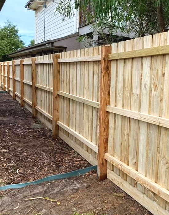 A wooden privacy fence under construction along a backyard, with a house and trees in the background.