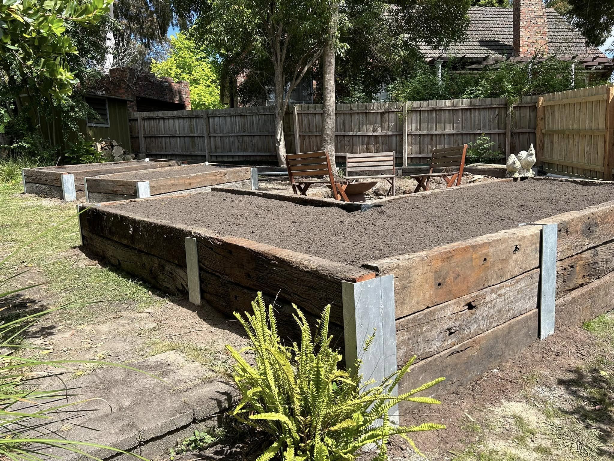 A backyard garden with raised wooden planting beds made from sleepers, a wooden bench, trees, and a wooden fence.