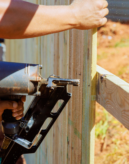 Person using a nail gun to attach wooden planks to a fence or structure outdoors.