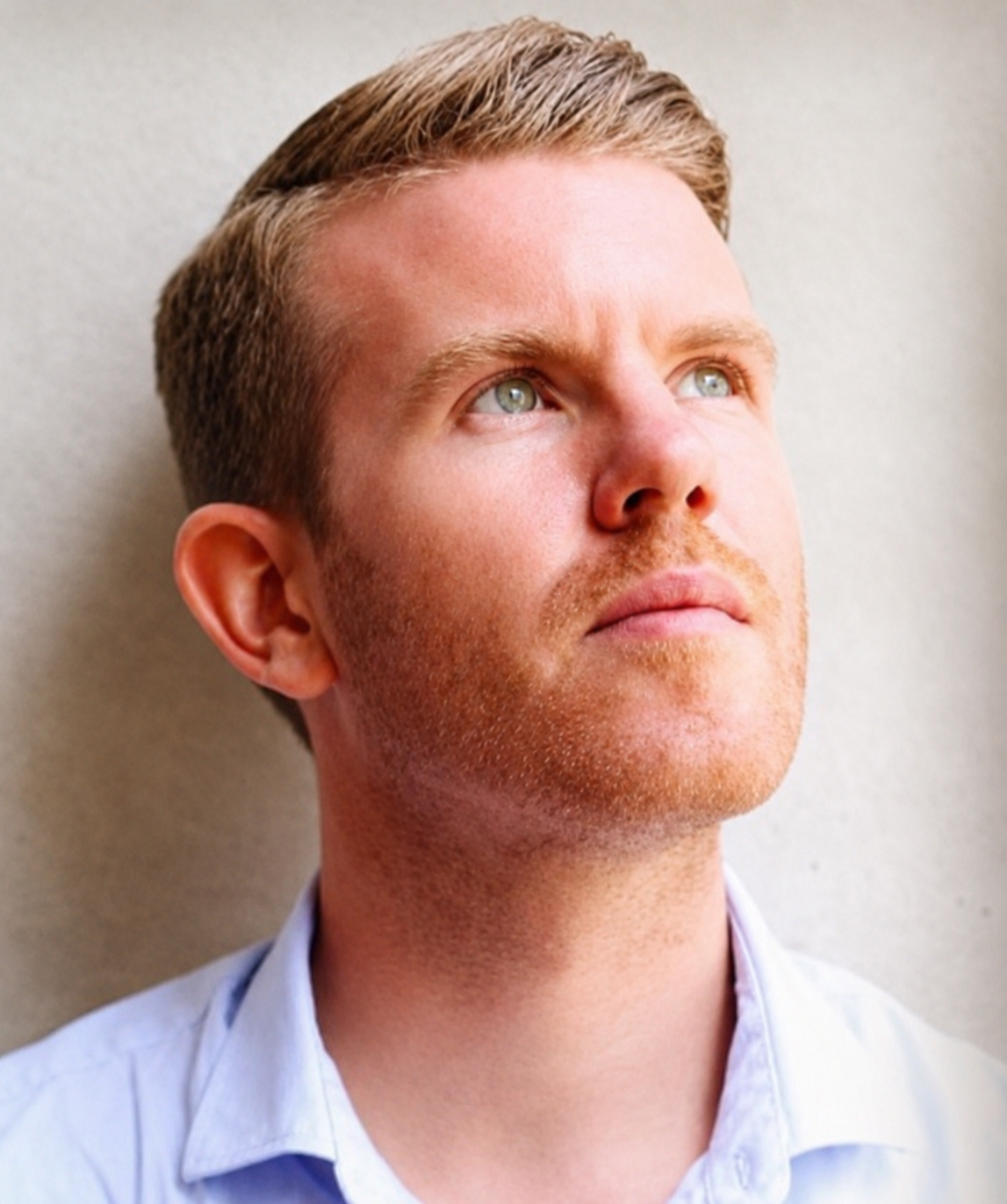 Close-up of a Sam Grebert with short, light brown hair and blue eyes looking upwards, wearing a light blue collared shirt, against a neutral background.