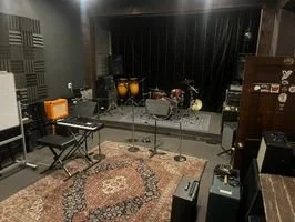 Music rehearsal room with drums, microphones, and amplifiers on a stage, and chairs and equipment in the foreground.