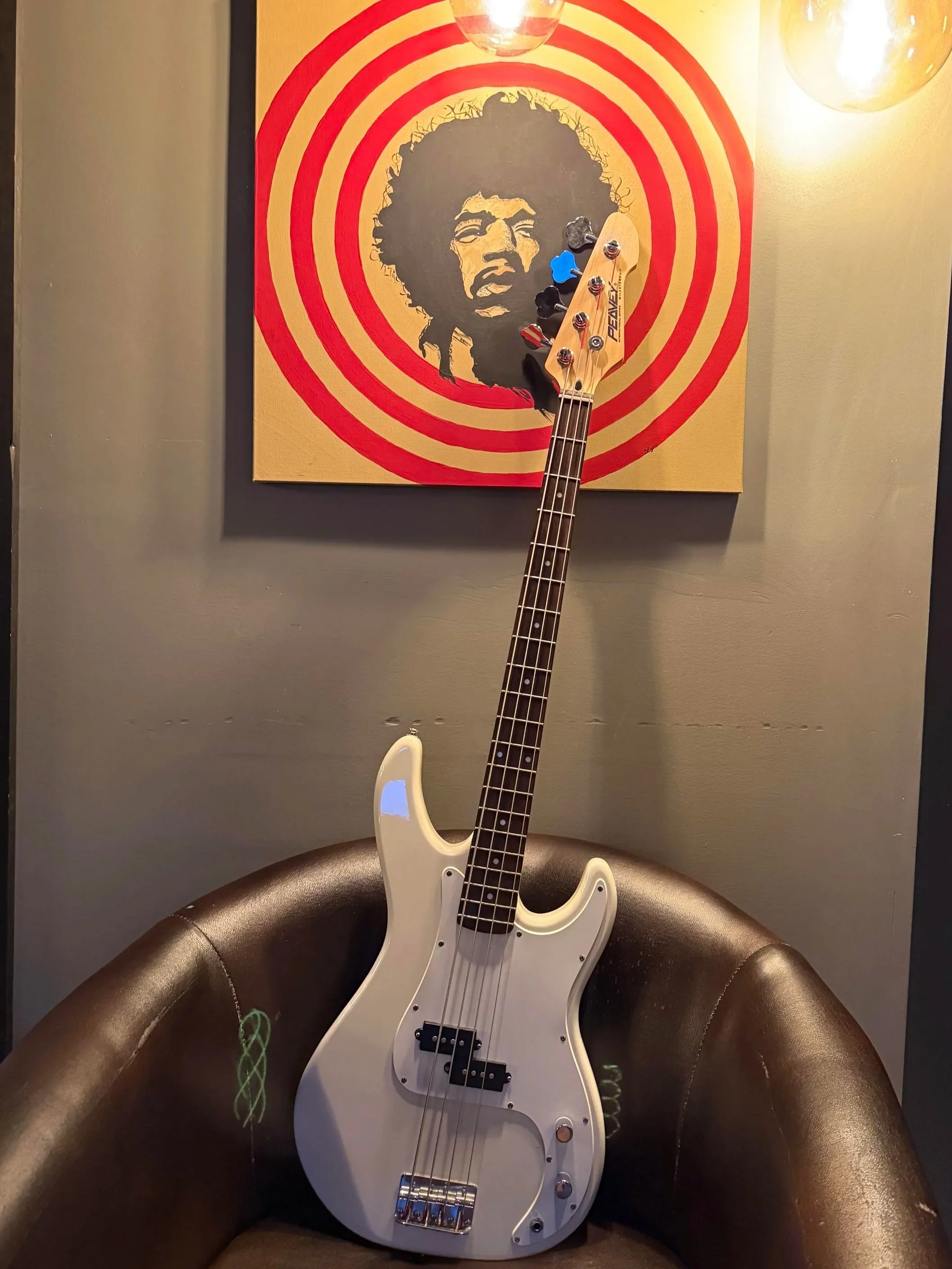 A white electric bass guitar resting on a brown leather chair in front of a wall with a portrait of a person with curly hair surrounded by red concentric circles.