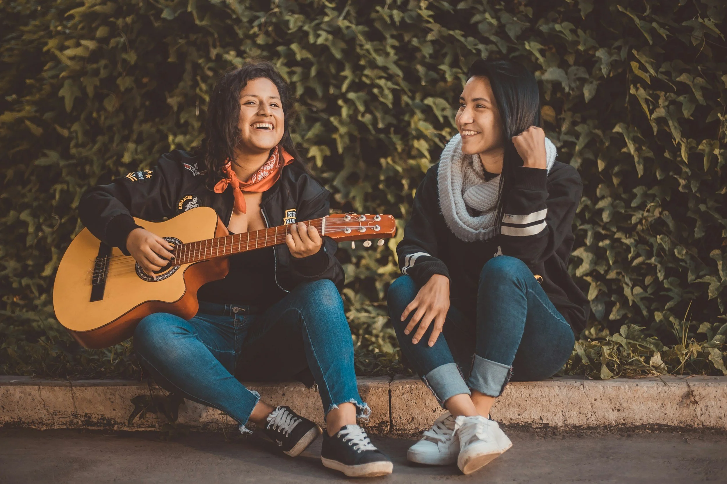 Two young women sitting on a low brick ledge in front of a bush of green ivy, smiling and enjoying each other's company. One woman is playing a guitar, while the other listens and smiles. They are casually dressed, and the scene appears to be outdoors in natural lighting.