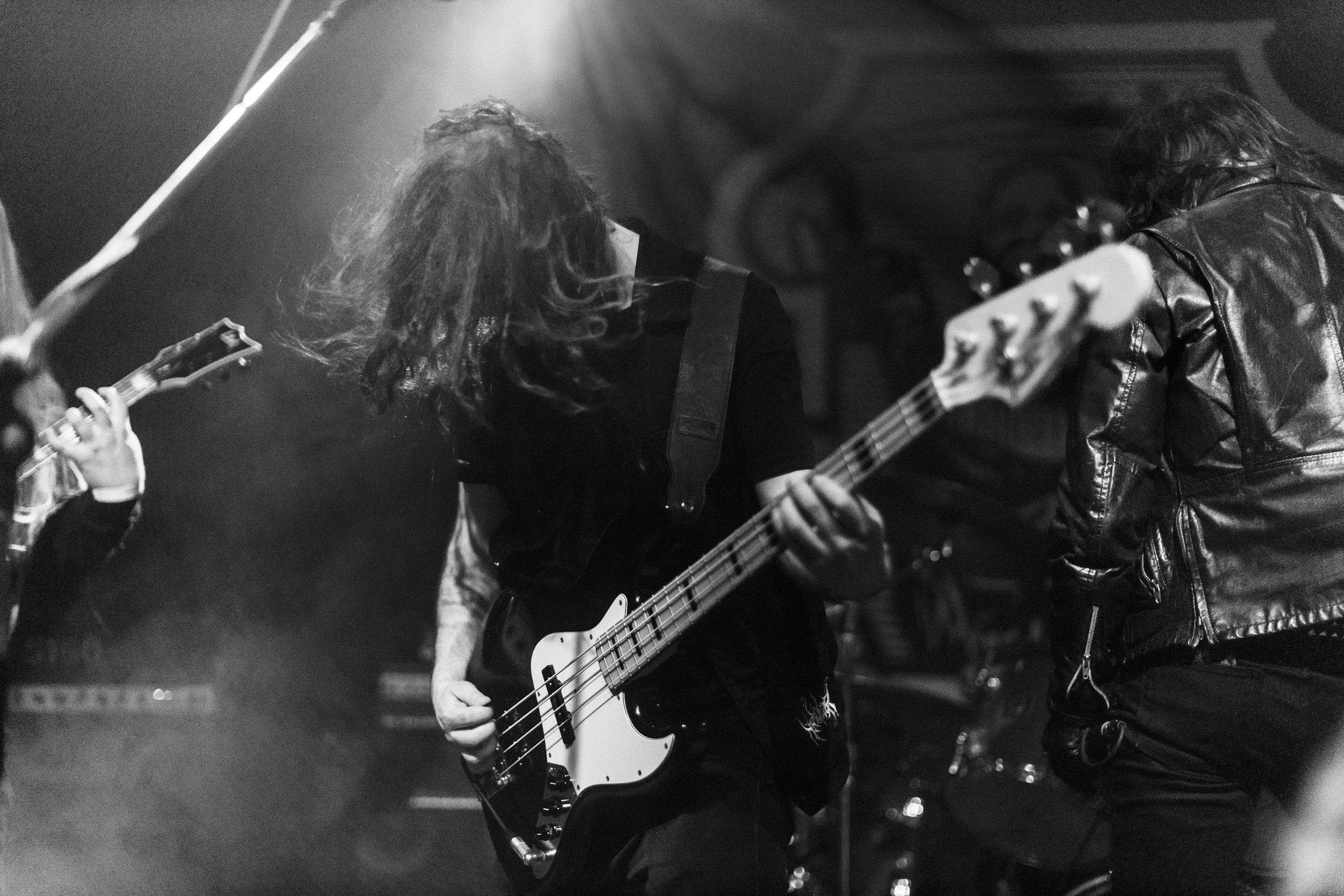 Black and white photo of two musicians playing guitars on stage, one with long, curly hair and the other with wavy hair and a leather jacket, in a dimly lit setting.