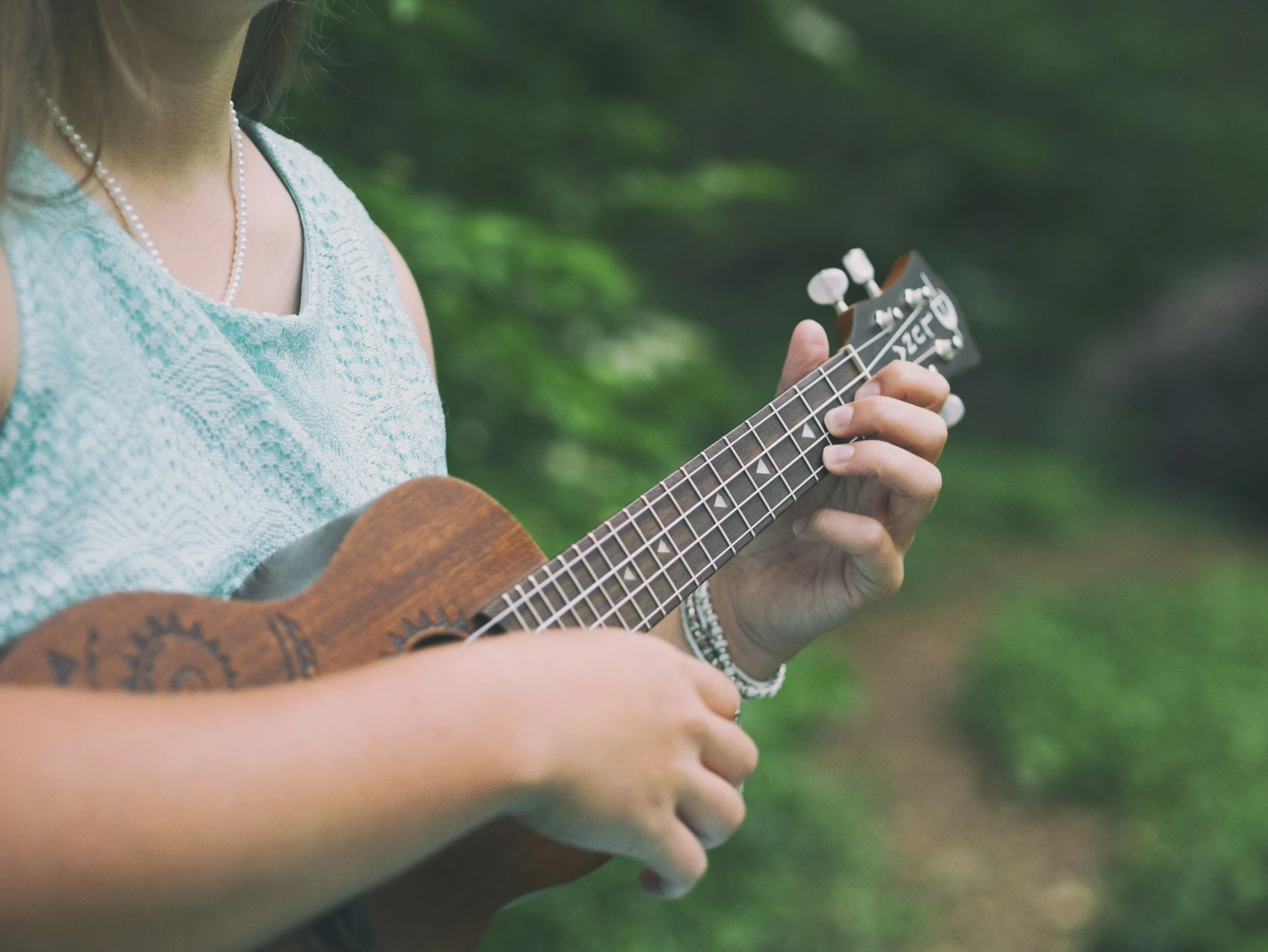 A woman in a light blue lace dress playing a ukulele outdoors with a blurred green background.