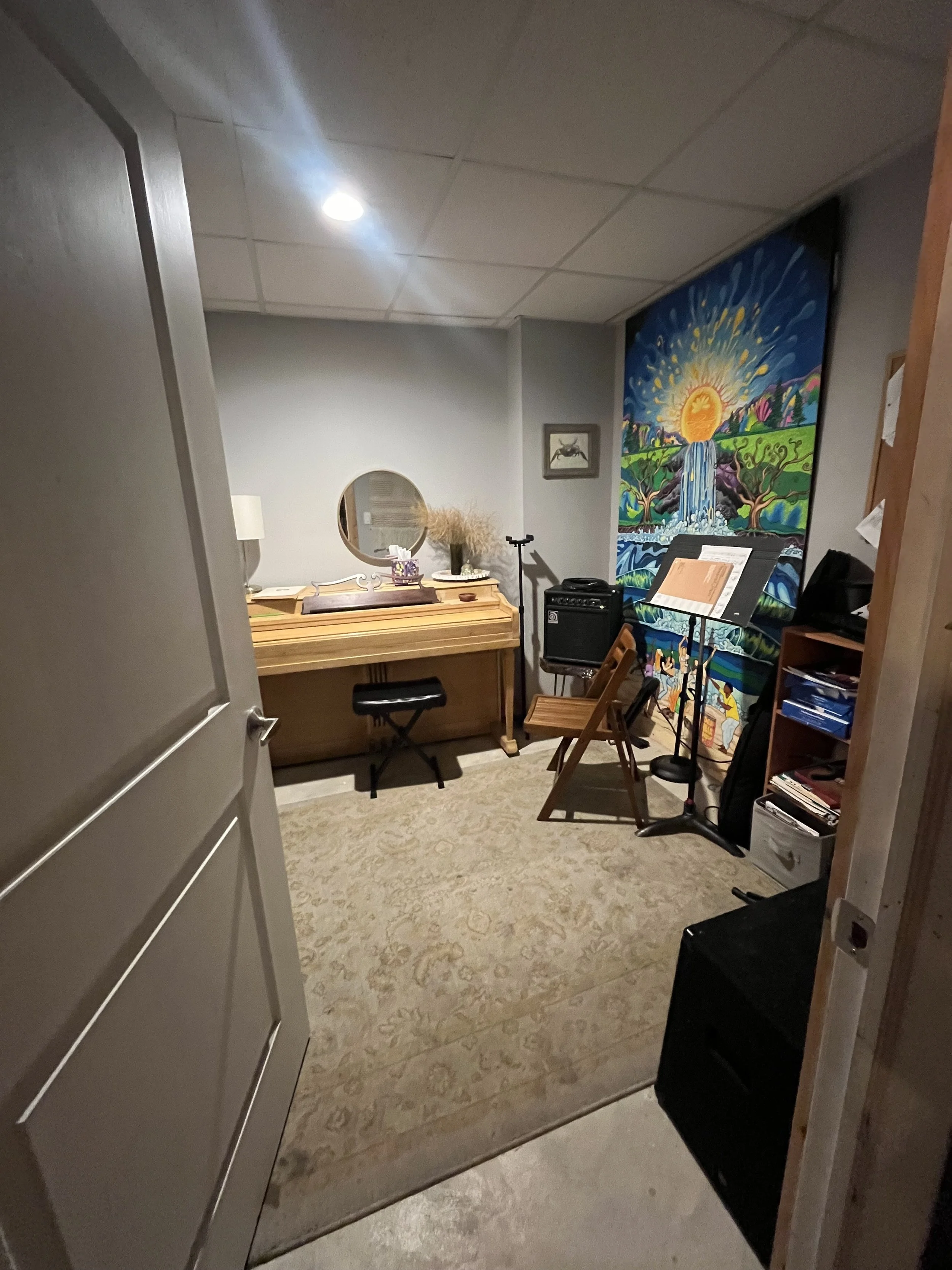 A cozy room with a small wooden upright piano with a round mirror and decorative items on top, a modern LED floor lamp, a colorful landscape painting on the wall, a music stand, and a folding wooden chair.