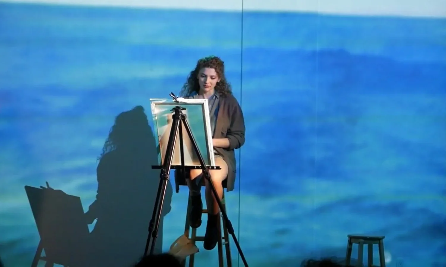 A vocal instructor sitting on a stool in front of a blue ocean-themed backdrop, with an easel and paintings from their theater performance.