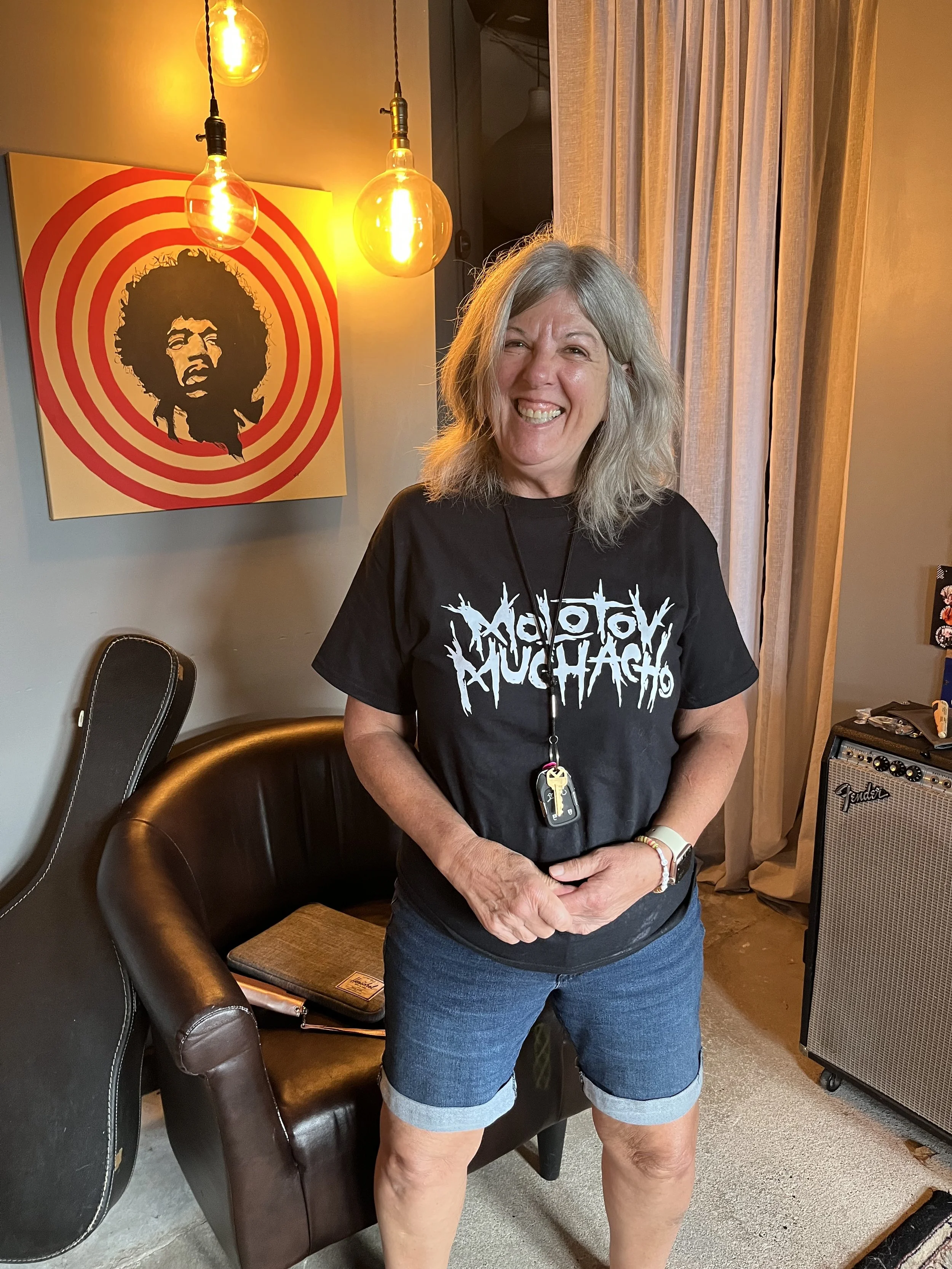 Smiling woman with gray hair wearing a black T-shirt with white text, standing in a room with beige curtains and a gradient guitar amplifier. Behind her is a framed artwork of a person's face with curly hair inside red and white concentric circles, accompanied by hanging Edison-style light bulbs.