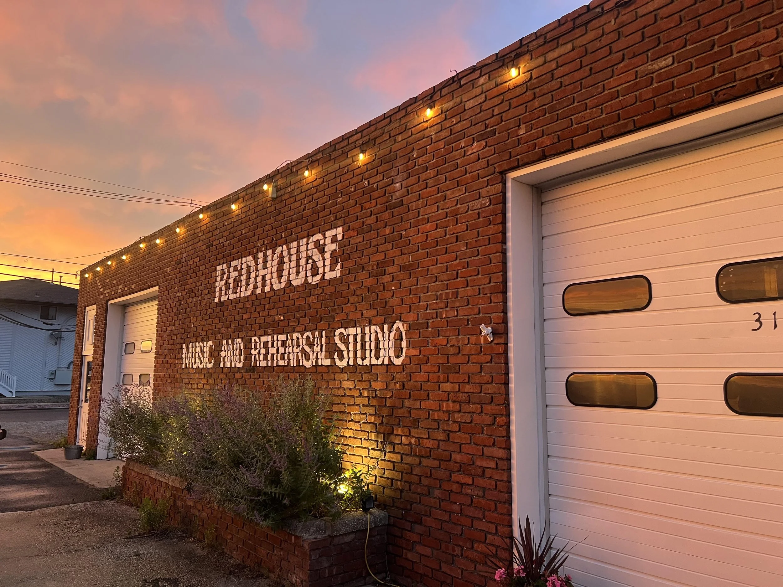 A brick building with a white garage door and a painted sign that reads 'FIREHOUSE' partially visible, with a sky at sunset in the background. String lights are hanging along the top of the wall, and there are plants and flowers at the base of the building.