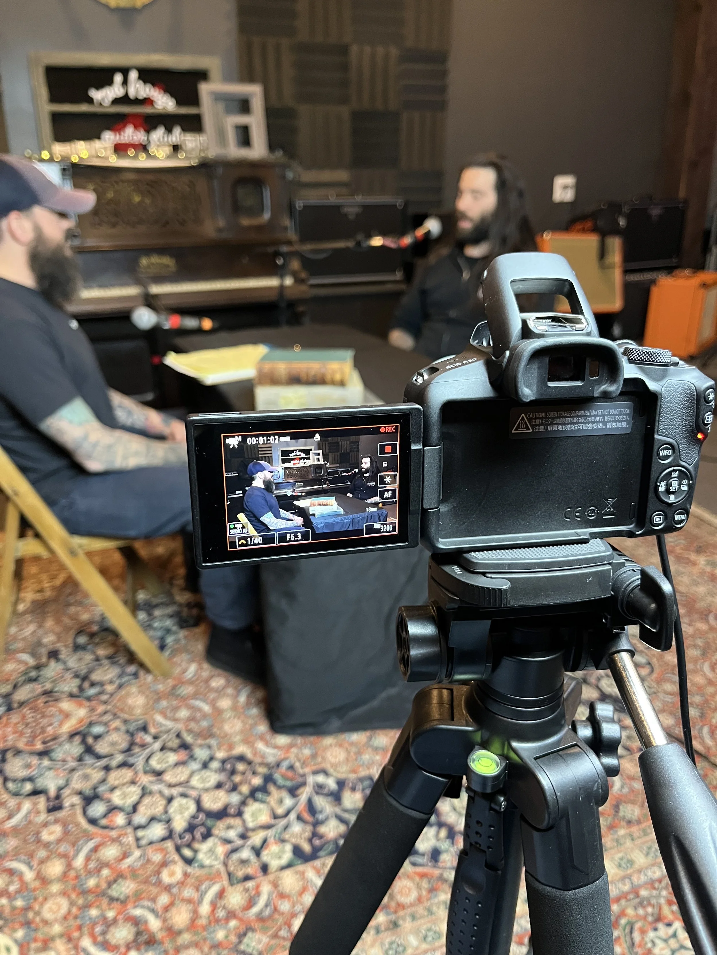 A camera on a tripod filming a music interview session in a studio, with two men talking in front of a piano.