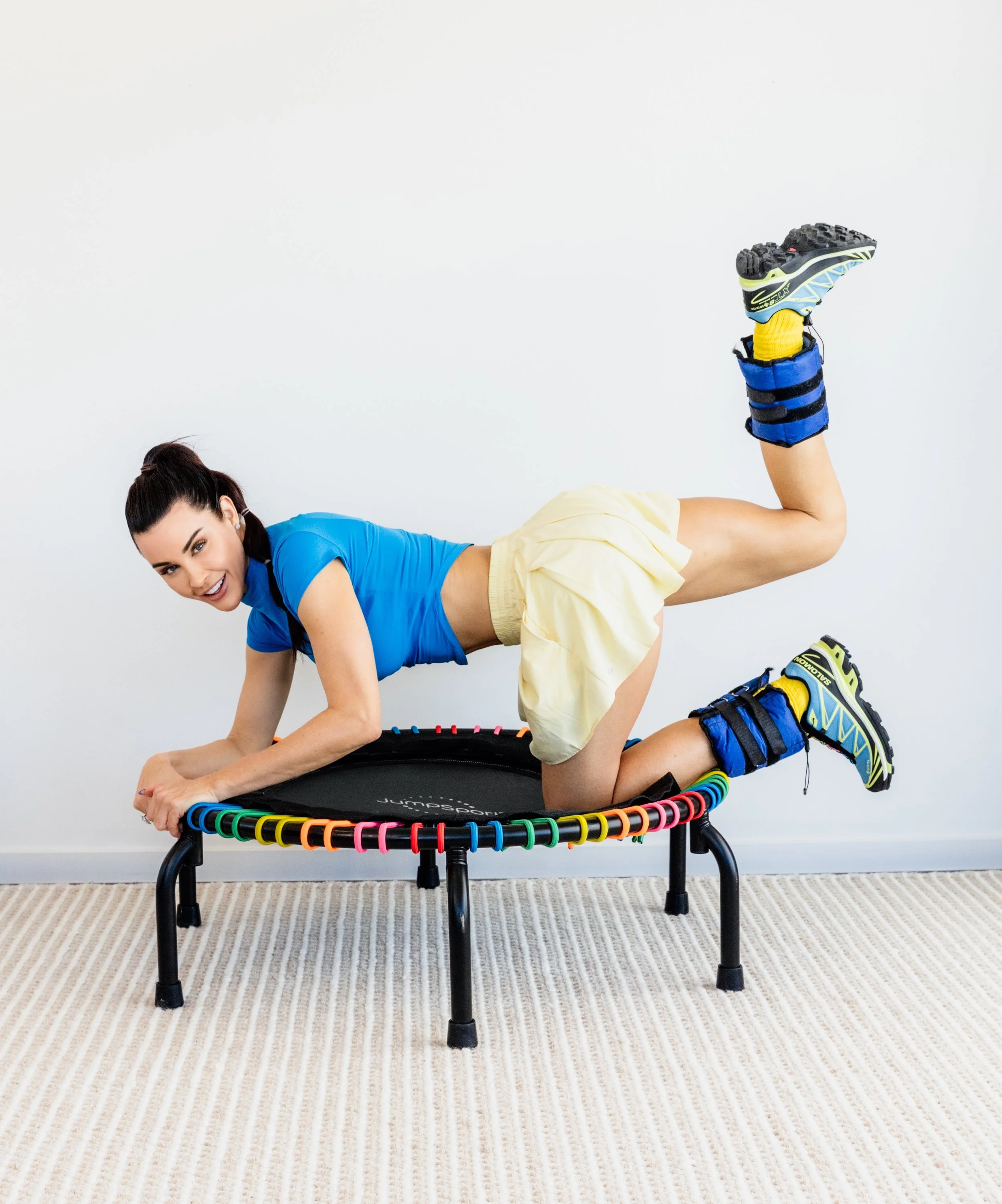 Woman lying on her side on a small trampoline, smiling, with her leg extended upward. She is wearing a blue shirt, yellow shorts, and colorful sneakers. The background is plain white, and the trampoline is on a beige striped carpet.
