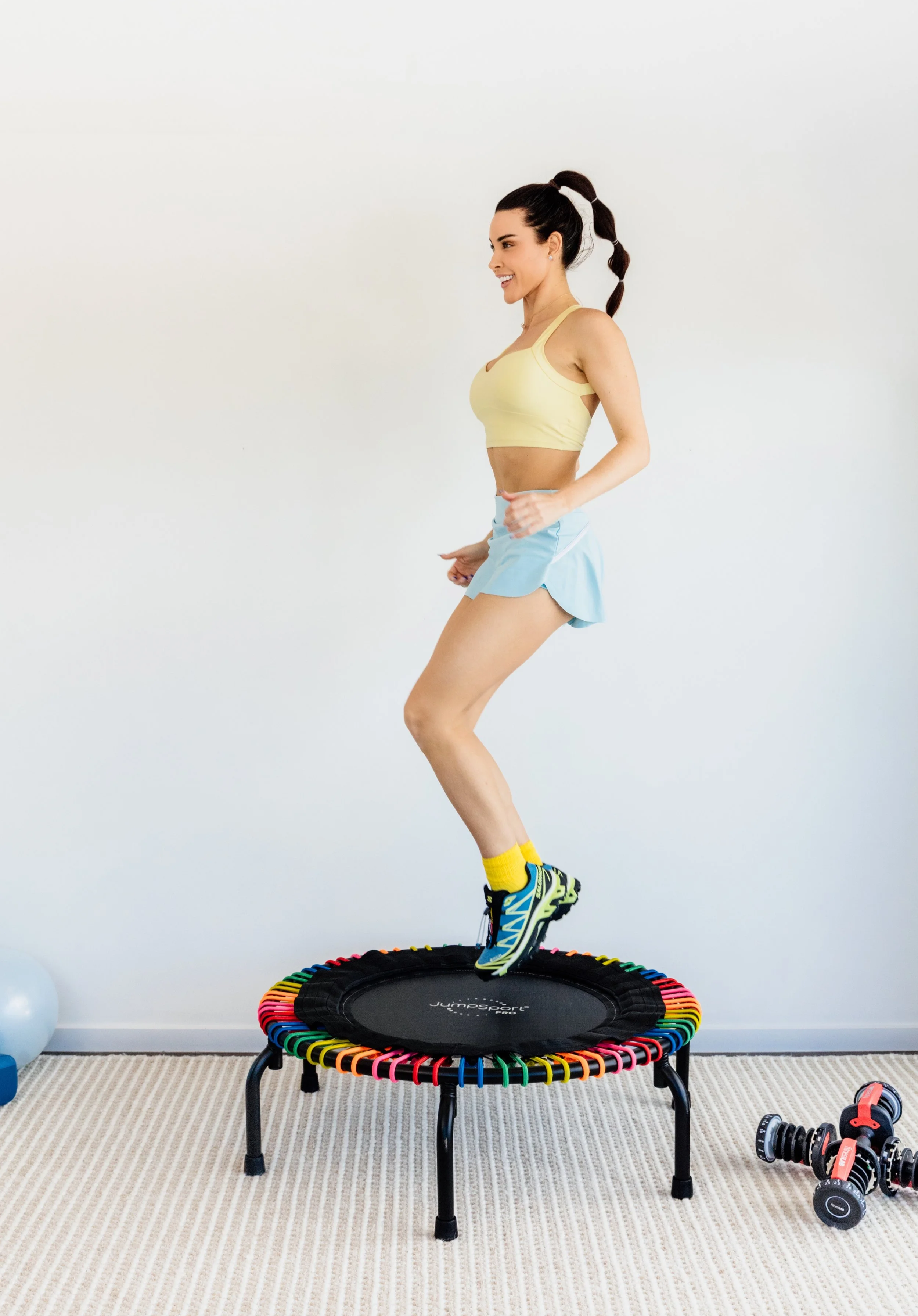 Woman jumping on a mini trampoline with a multicolored border in a room with a striped beige carpet, exercise equipment including dumbbells, and a white wall.