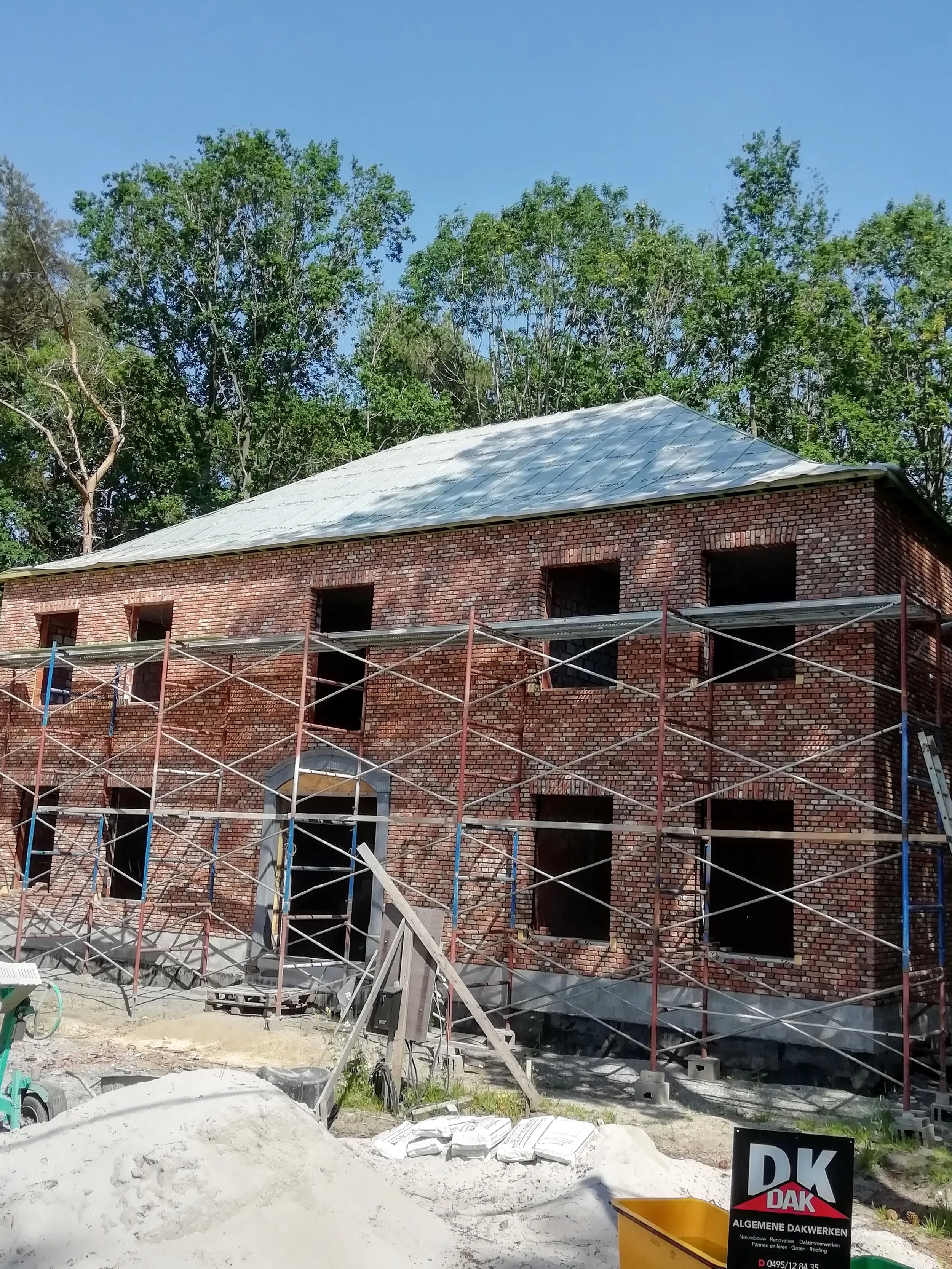 Een bouwproject met een twee verdiepingen tellend gebouw in aanbouw, met een nog niet afgewerkte bakstenen gevel, omlijst door groen bomen en een blauwe hemel. Er staat scaffolding rondom het gebouw en er liggen bouwmaterialen op de grond.