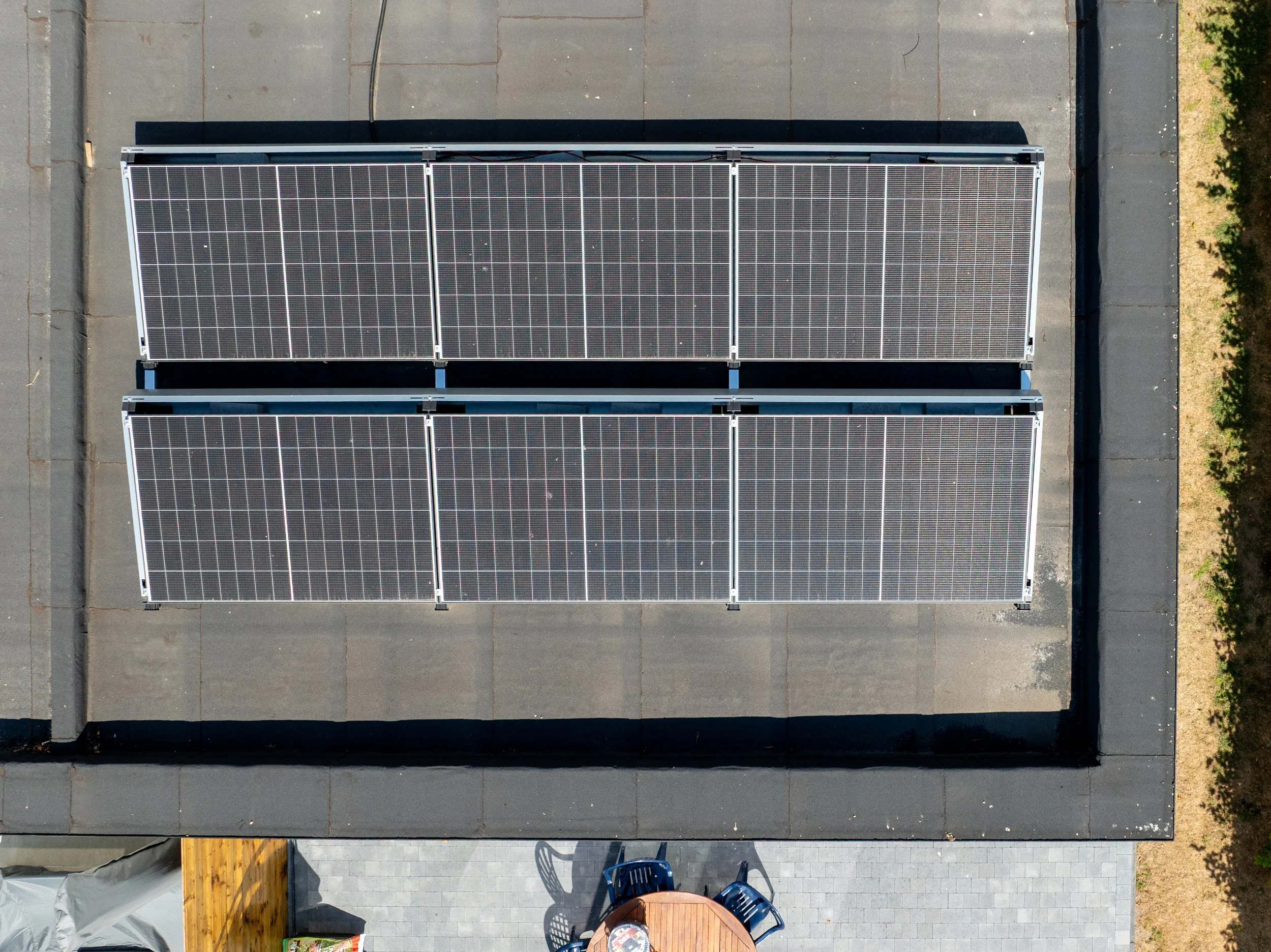 Bovenaanzicht van een dak met zonnepanelen. Onder aanzicht van een terras met een ronde tafel en vier blauwe stoelen.