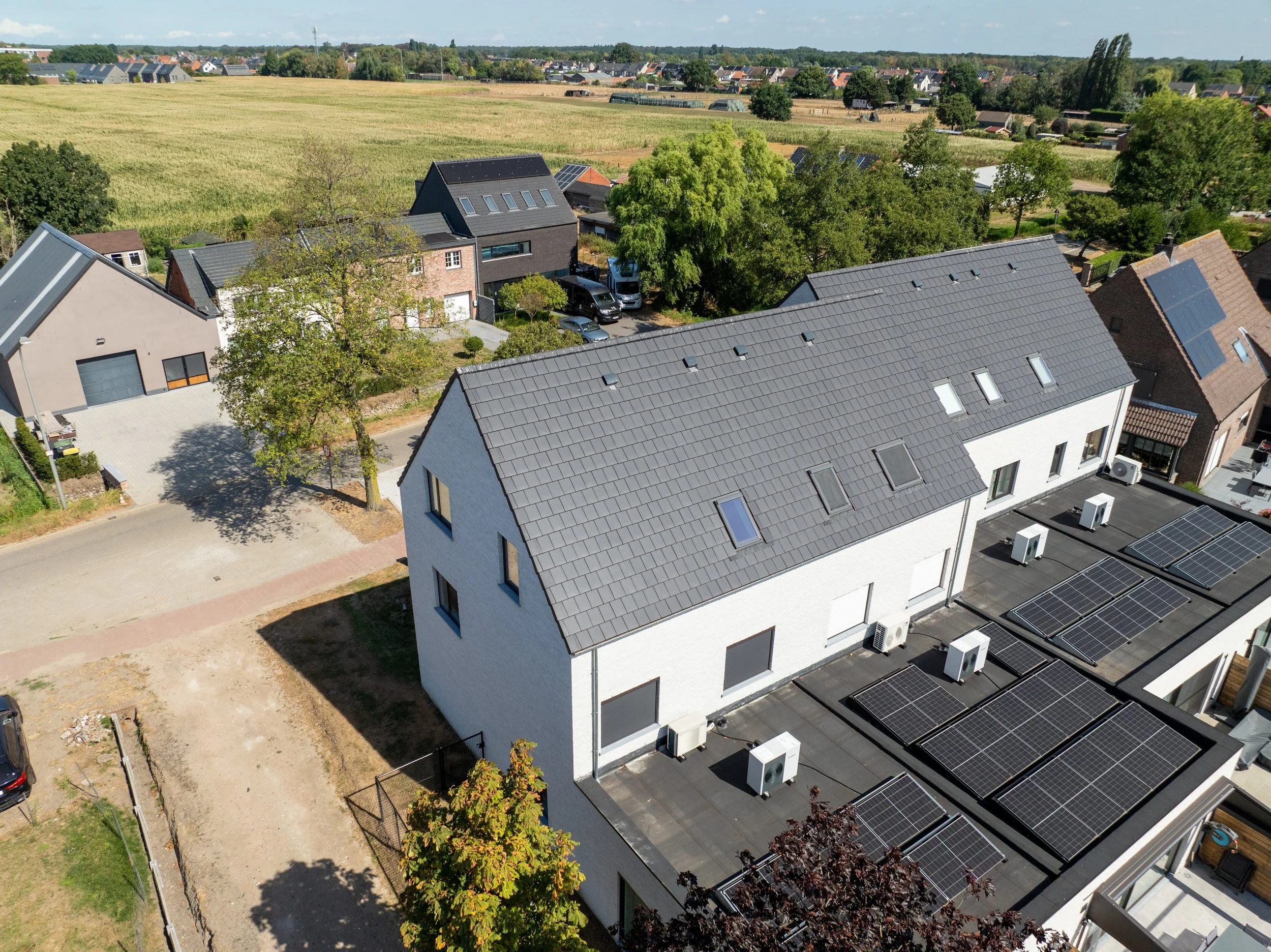 Luchtfoto van een residentiële wijk met huizen en daken, sommige met zonnepanelen, tussen groene bomen, op de achtergrond velden en een dorp.
