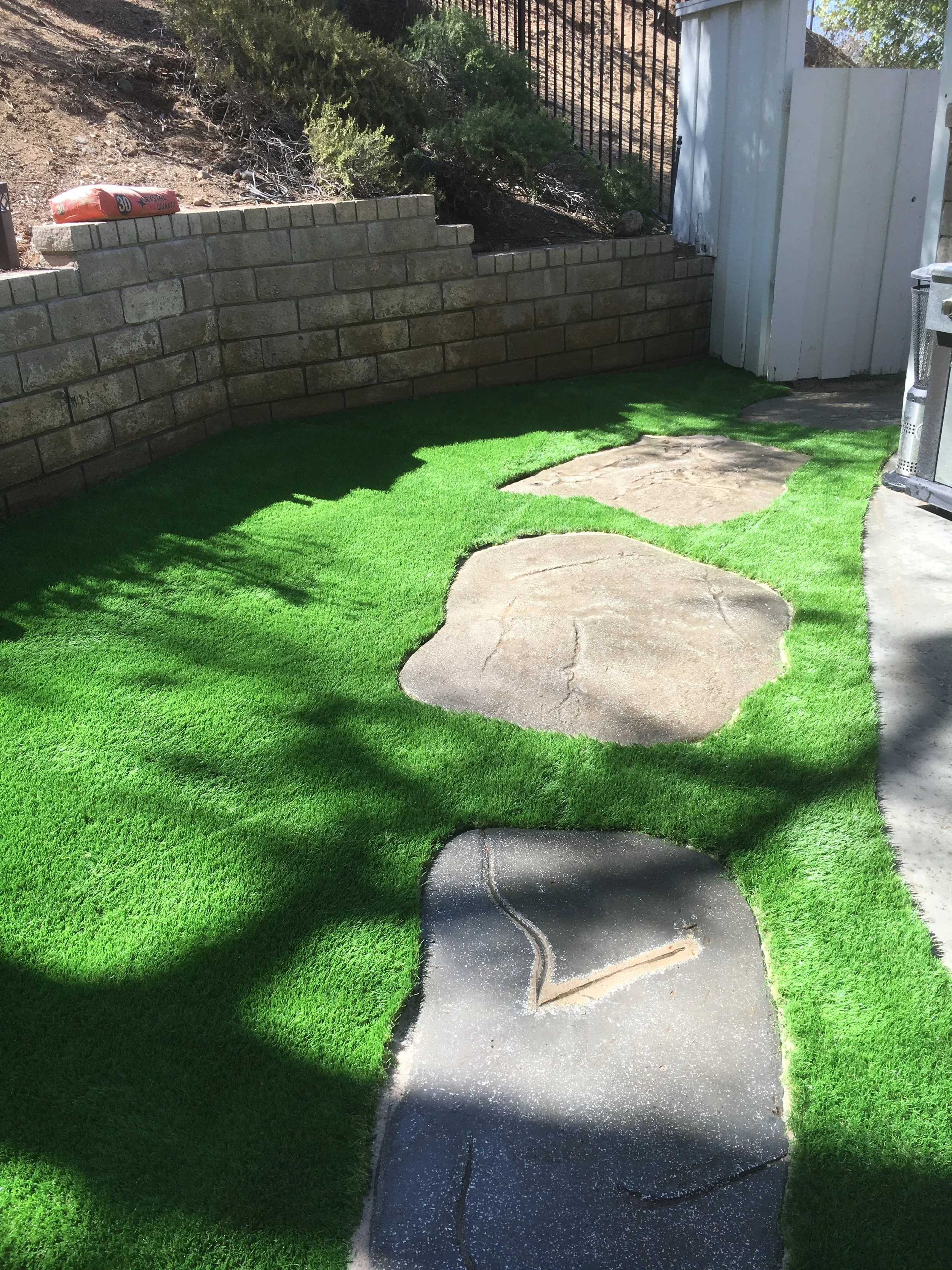 A backyard scene showing a concrete pathway with cracks, surrounded by vibrant green artificial turf, a stone wall with a raised garden bed behind it, and a white wooden fence on the right. There is a clear sunny sky casting shadows.