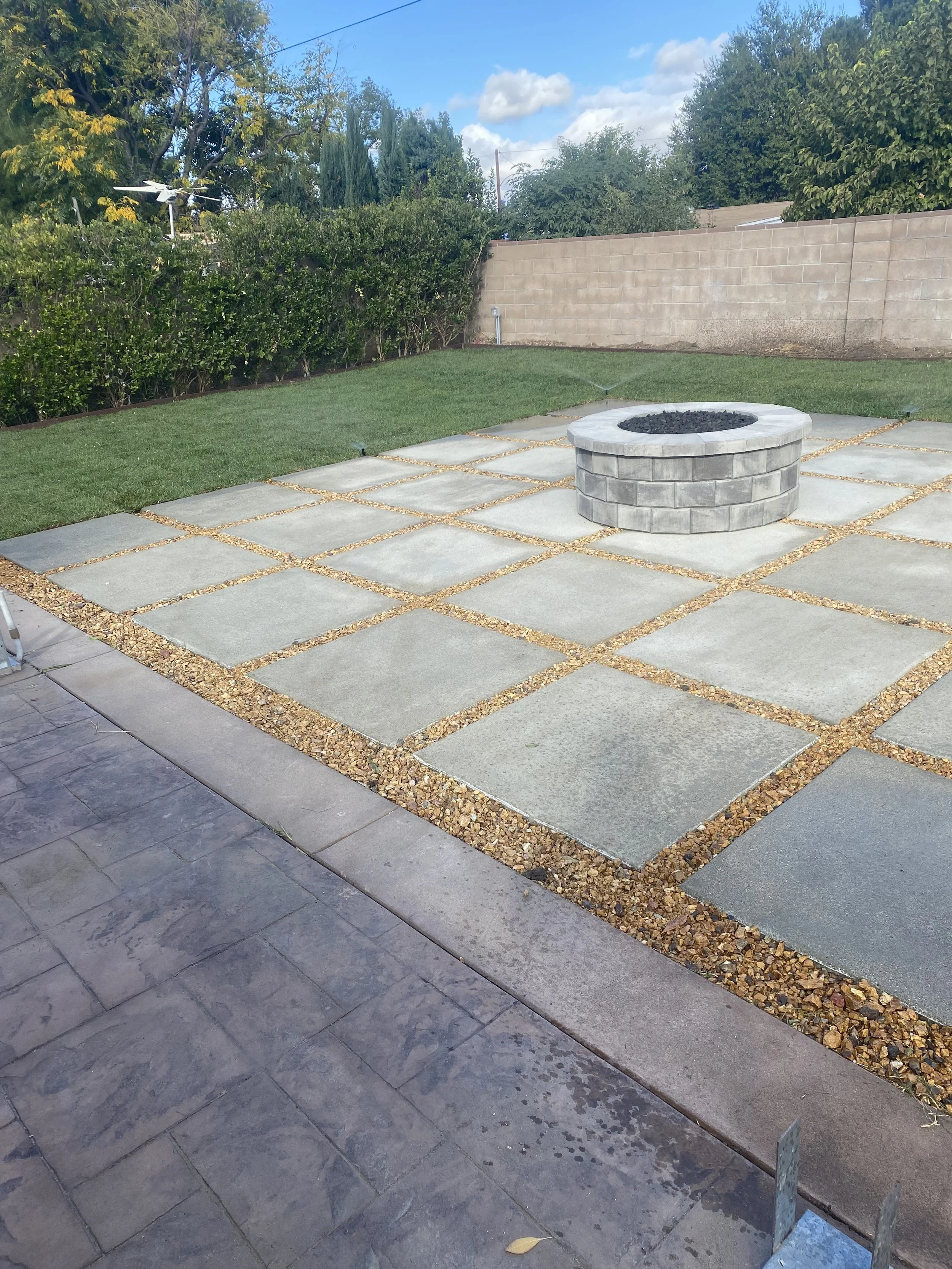 A backyard with a paved patio area with square concrete slabs separated by small gravel strips. There is a round fire pit in the center made of gray bricks filled with black gravel. Behind the patio, there is a strip of grass, a hedge and a beige brick wall. A white weather vane is visible over the hedge. The sky is partly cloudy with a few clouds and trees are visible over the wall.