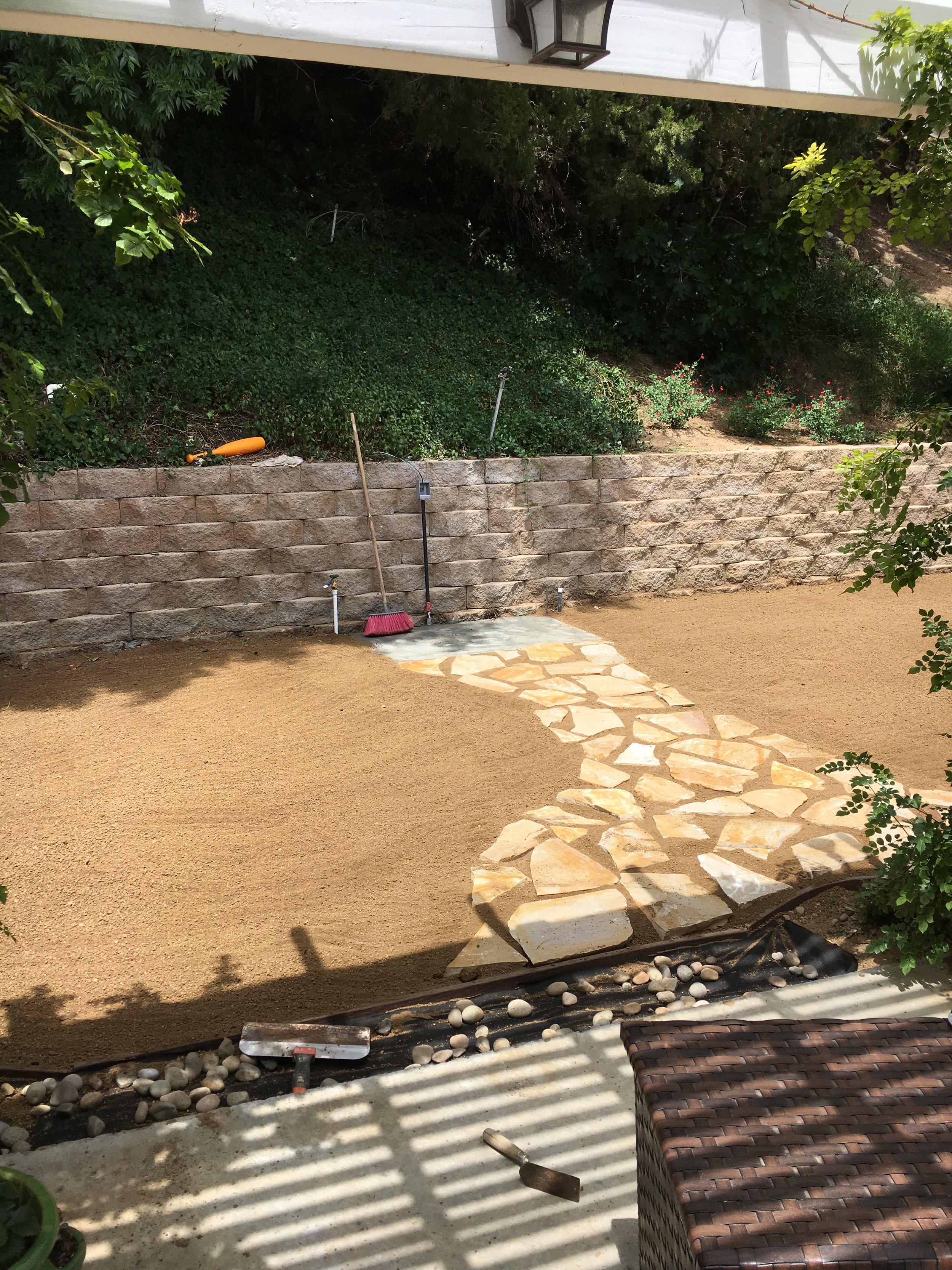 A backyard with a stone pathway leading to a brown wicker chair. Freshly laid sandy ground surrounds the pathway, with a stone retaining wall topped with green bushes and trees. Gardening tools like a broom, a small level, and a trowel are visible along the wall, with a flower bed containing small flowering plants to the right.