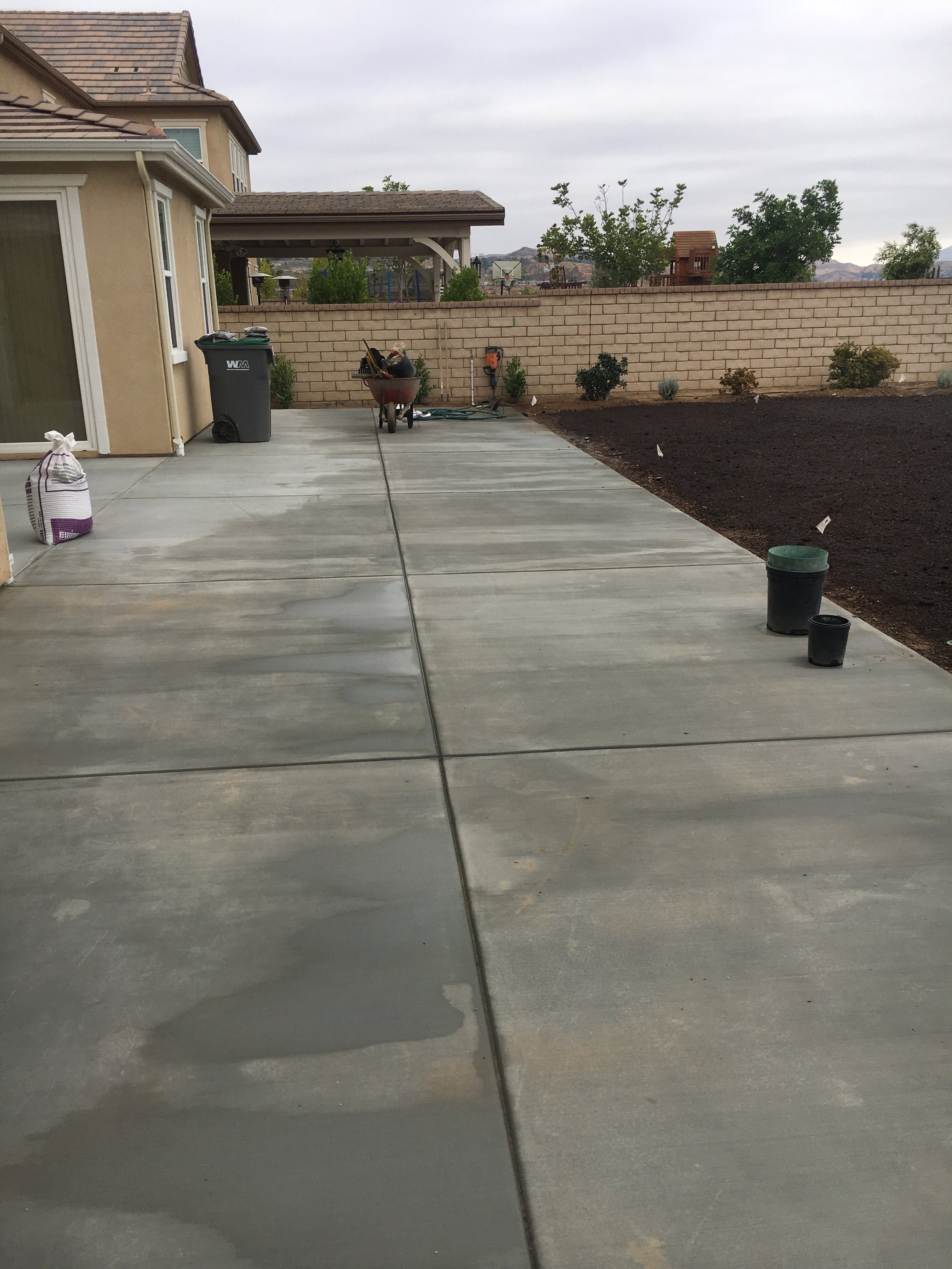 Backyard with a concrete patio, a wheelbarrow filled with tools, a trash bin, a garden bed with plants, and a brick wall with a neighborhood and hills in the background.