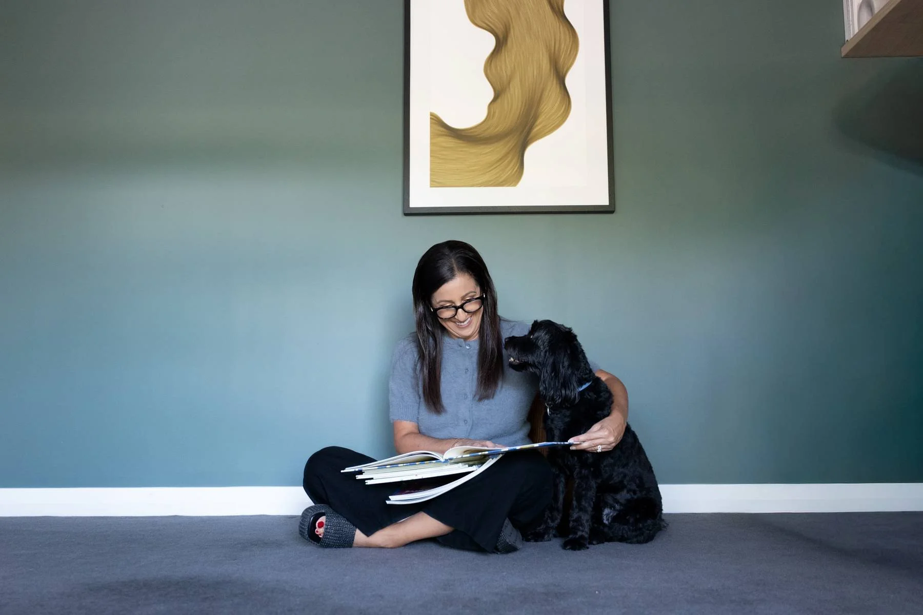 A woman sitting on the floor reading a book while a black dog sits beside her, looking up at her. The woman is smiling and wearing glasses and gray shirt. The background is a green wall with a framed piece of art depicting blonde hair.