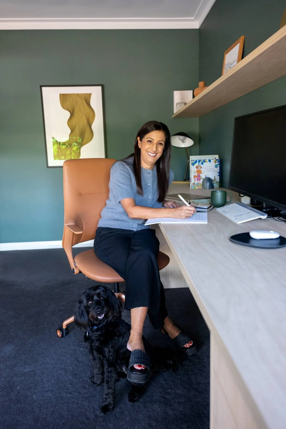 A woman sitting at a desk in a home office, smiling at the camera and writing in a notepad. There is a black dog sitting on the floor in front of her, looking up. The desk has a monitor, keyboard, mouse, and a green mug. The background features a green wall with artwork, shelves with decorative items, and a small table lamp.