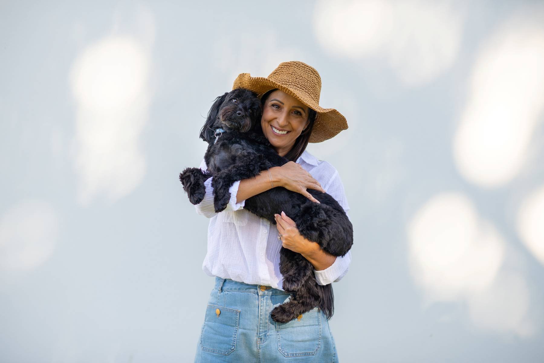 A woman in a white shirt and blue jeans, wearing a wide-brimmed straw hat, holding a black dog with curly fur. The woman is smiling and standing against a light background with bokeh light effects.