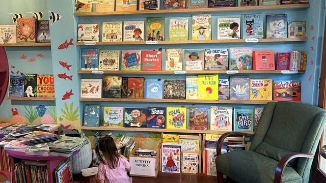 Library with bookshelves filled with children's books, a young girl sitting on the floor, and a green armchair near a window.
