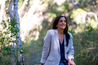 Woman smiling outdoors in a park with green foliage and trees in the background.