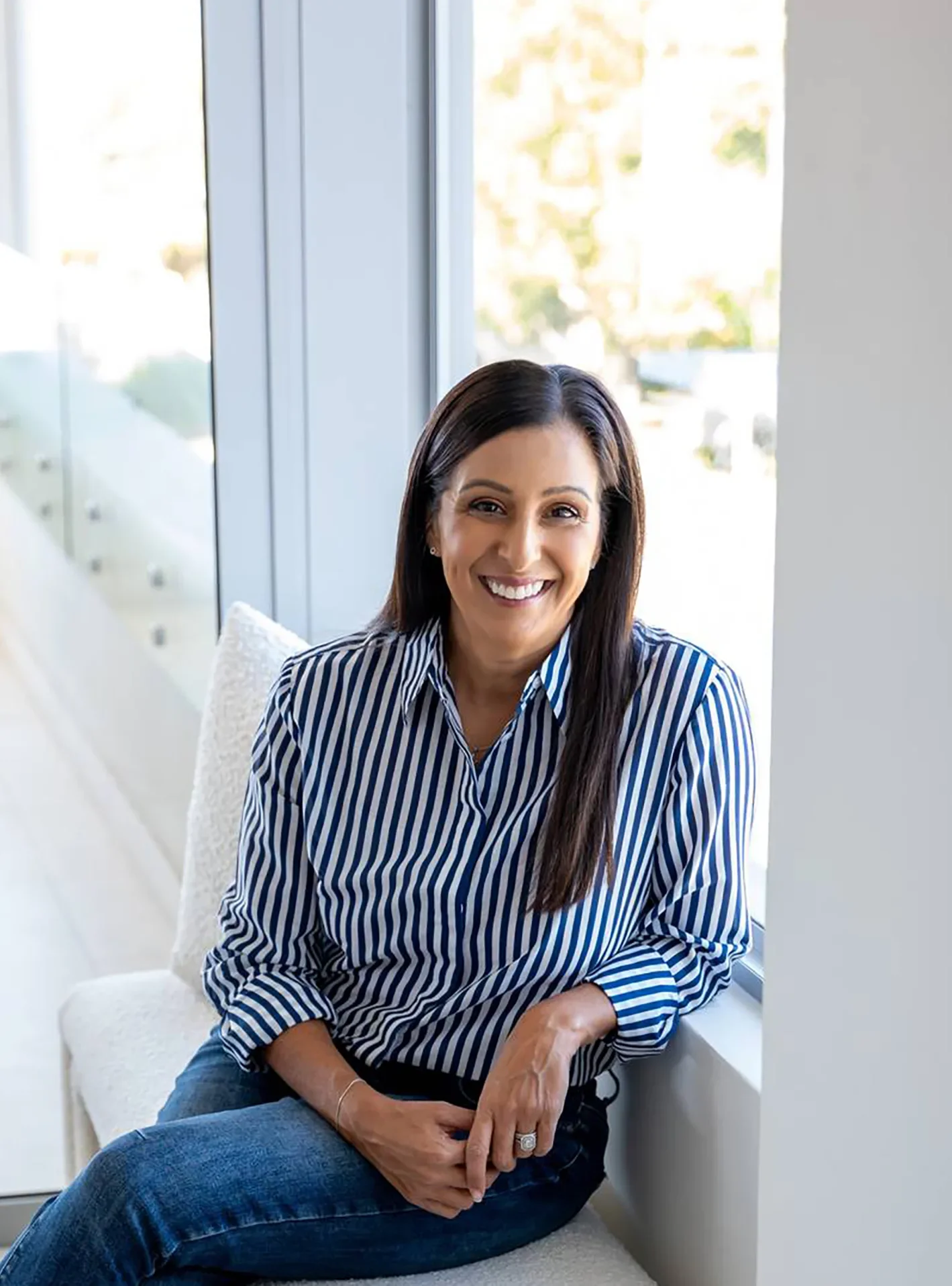 Woman with long dark hair smiling, sitting by a large window with a cityscape view, wearing a blue and white striped shirt and blue jeans.