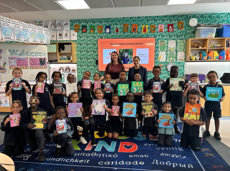 Group of young students holding books in a classroom with teachers behind them and educational decorations on the walls.