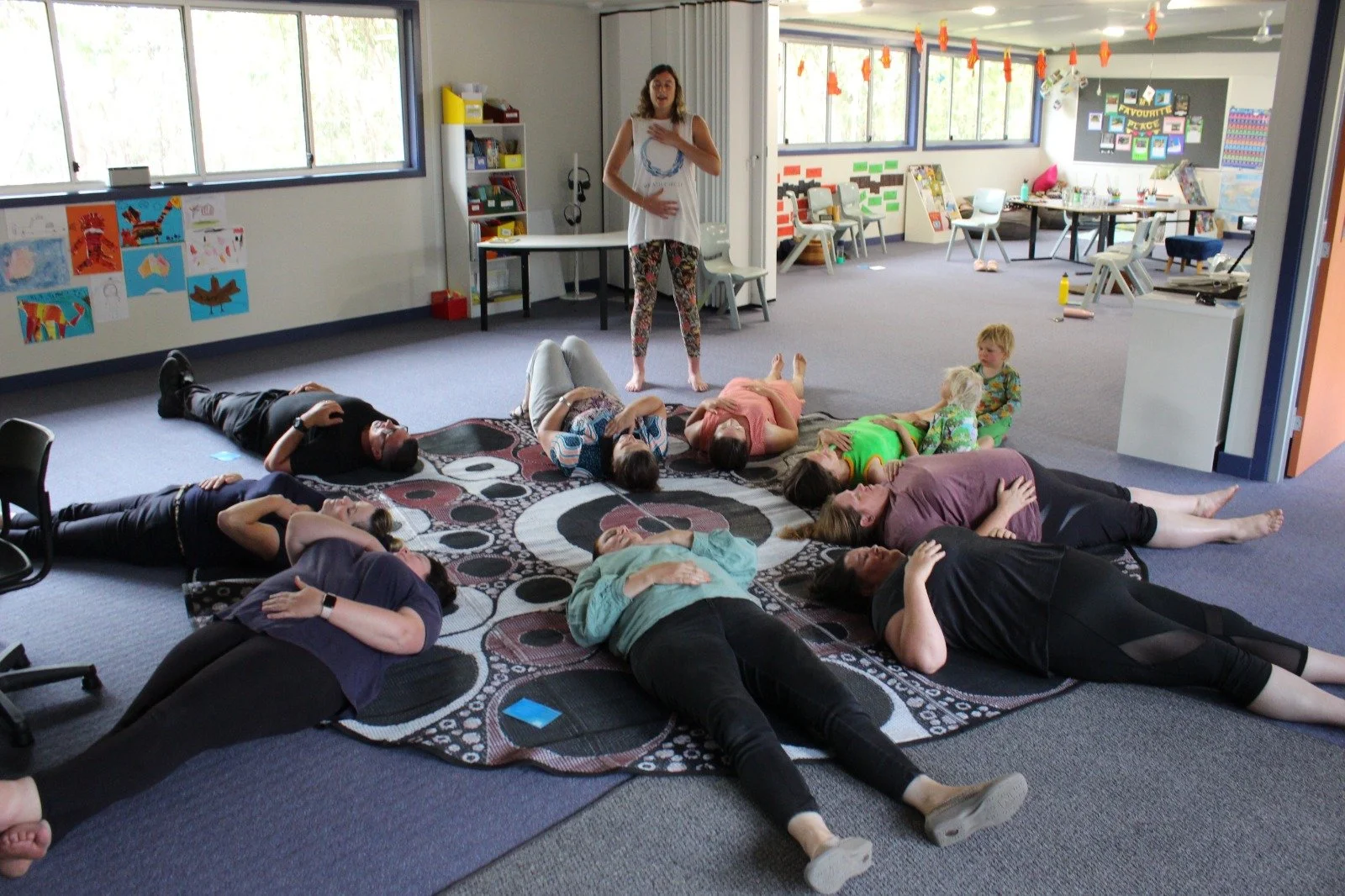 A group of adults and children lie on the floor on a patterned rug, participating in a relaxation or yoga session indoors. A woman stands at the front, leading the activity with her hand on her chest. The room has large windows, colorful artwork on the walls, and a counter with a bulletin board in the background.