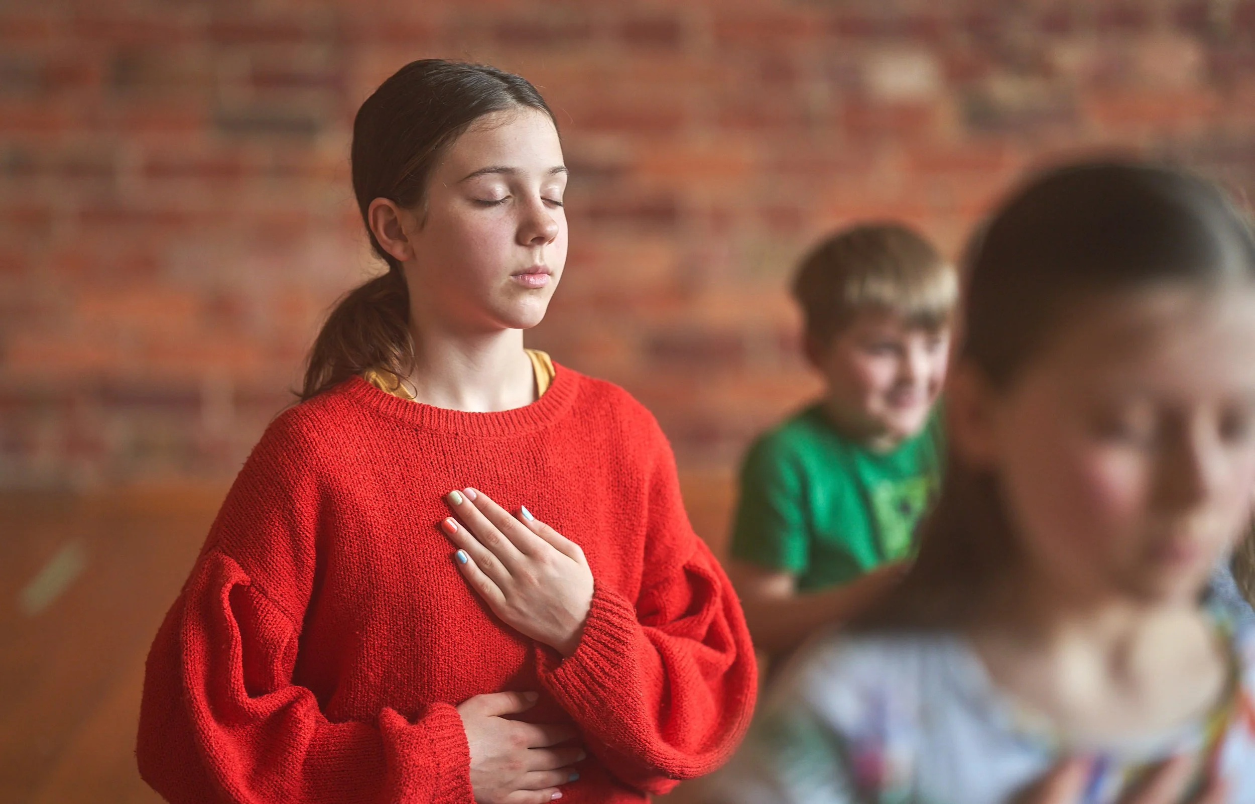 A girl with closed eyes and hand on her chest, wearing a red sweater, in a group of children who seem to be meditating or praying.
