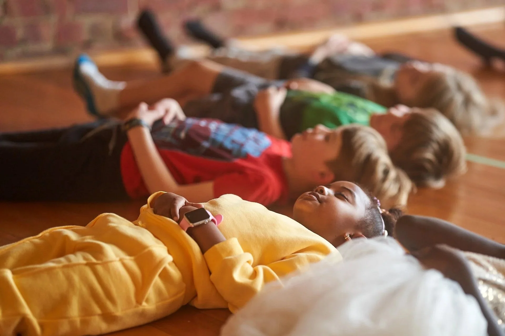 Children lying on a wooden floor, sleeping or resting, with their heads and bodies touching, in a room with exposed brick wall.
