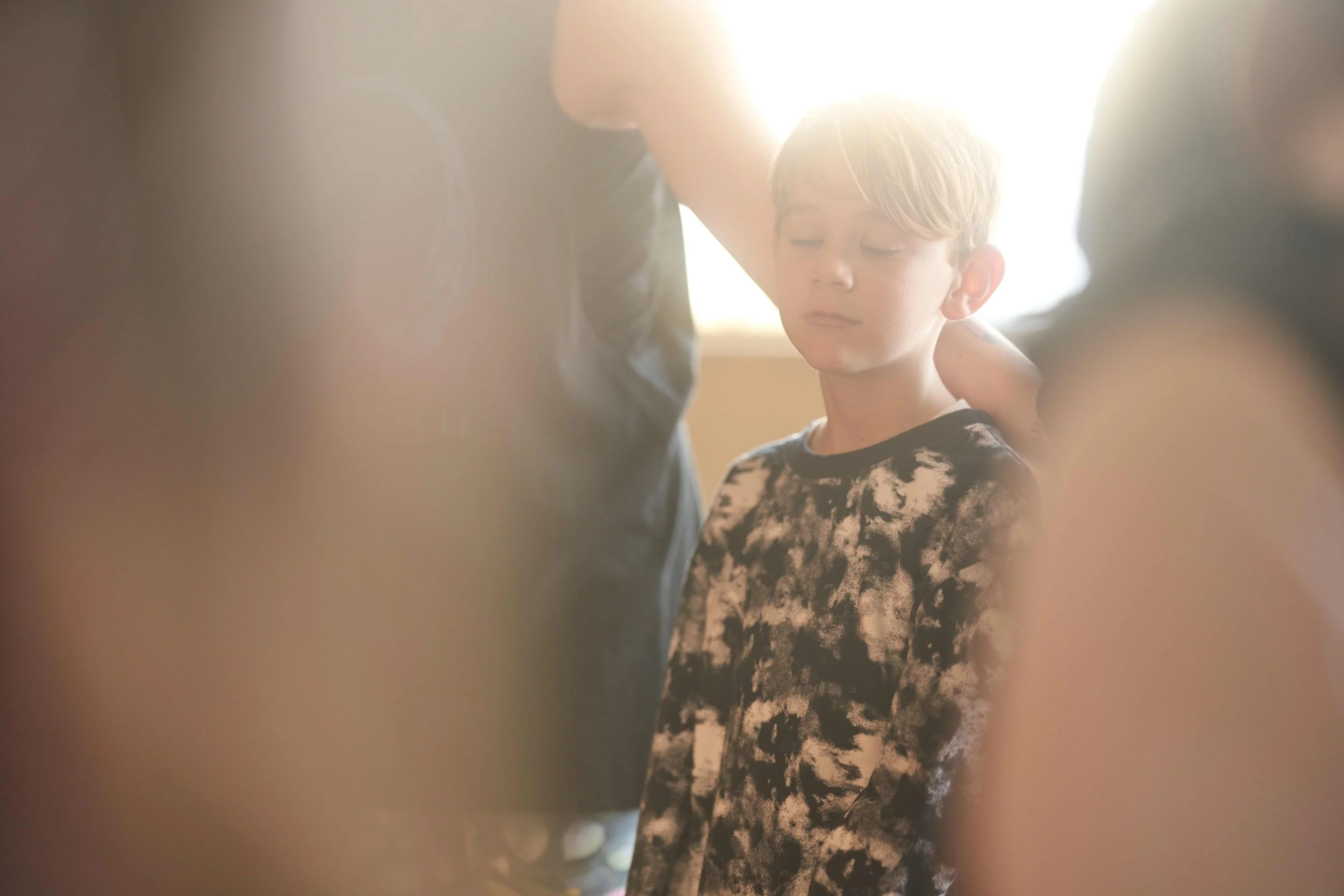 A young boy with short blonde hair and a patterned shirt has her eyes closed as a person adjusts her hair or head, with sunlight backlighting the scene.