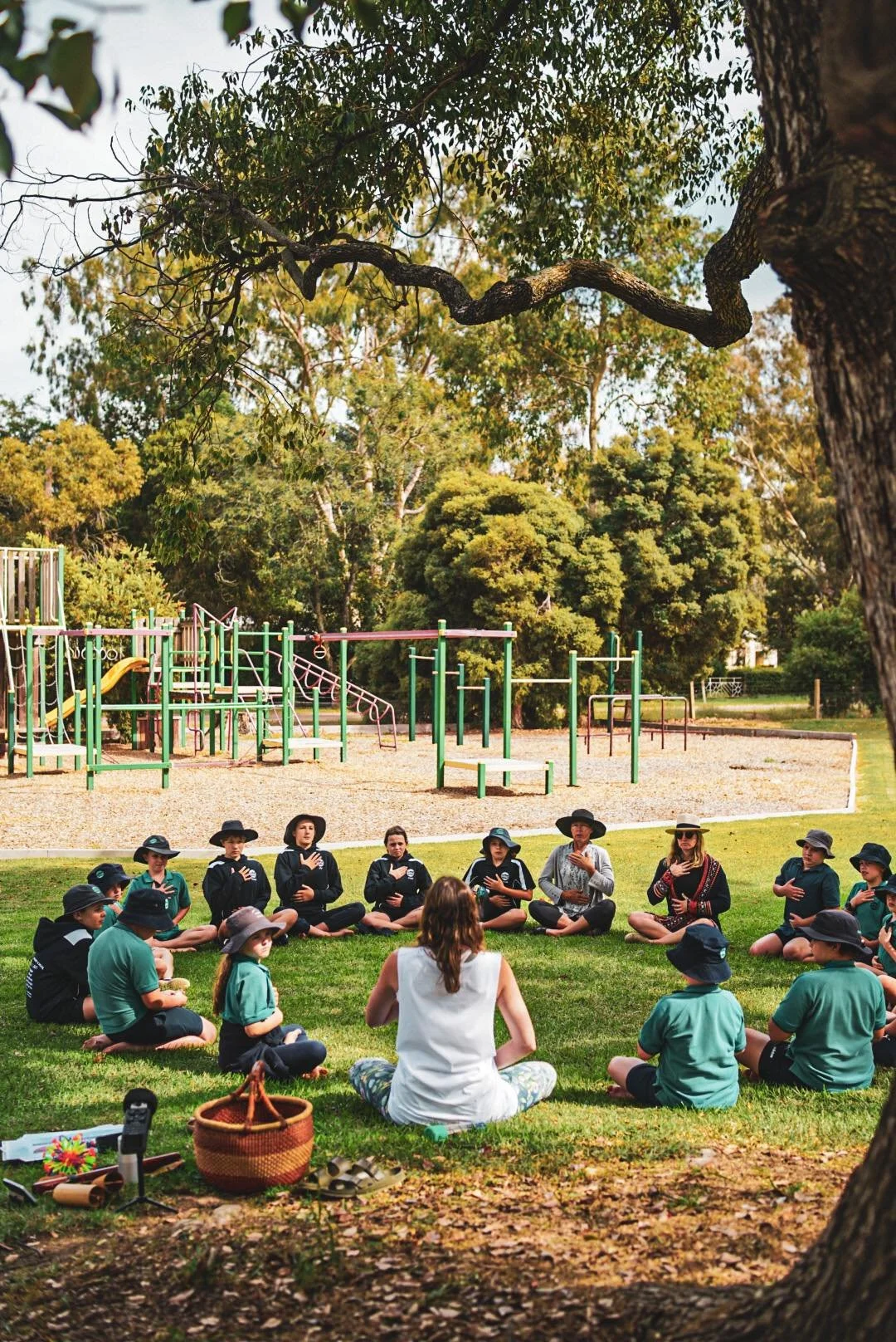 Group of children and adults sitting in a circle on a grassy field in a park, participating in an outdoor activity or discussion, with playground equipment and trees in the background.