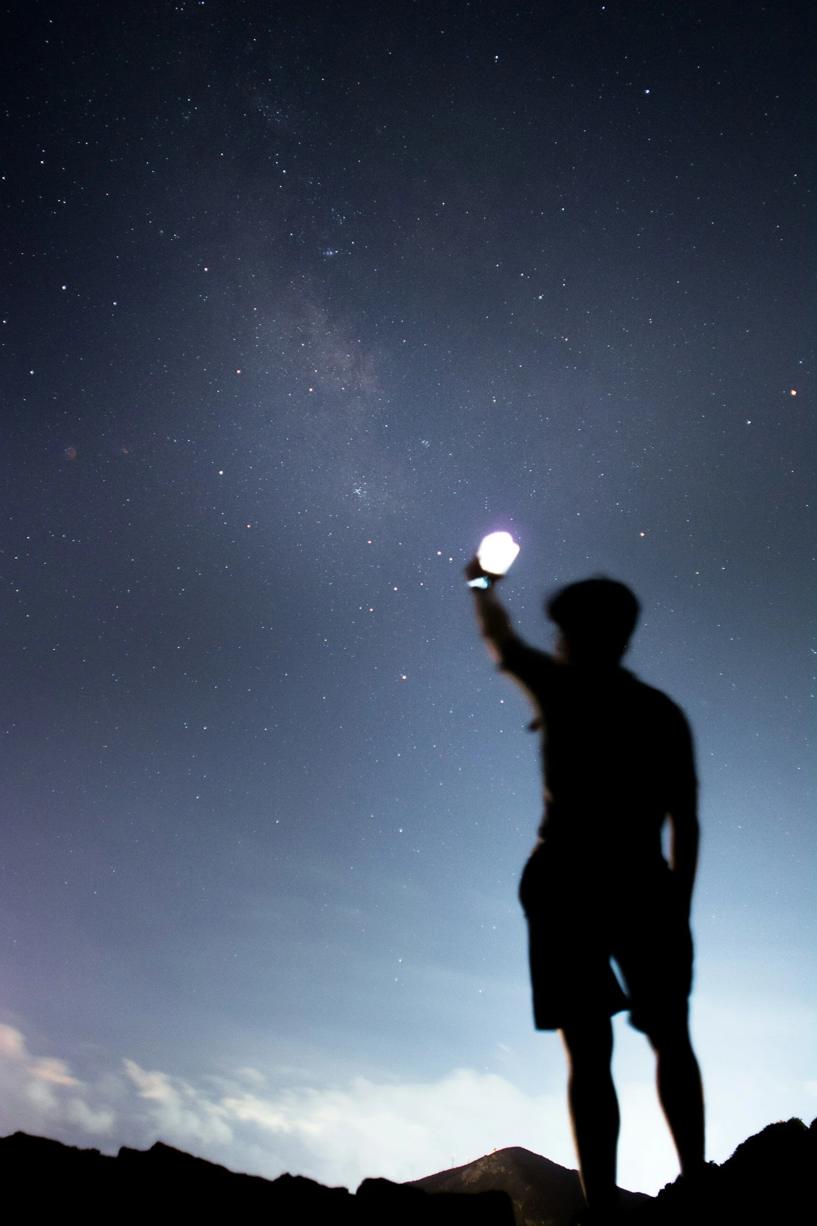 Person silhouetted against the night sky, holding a bright lantern or light, with stars and the Milky Way visible above.