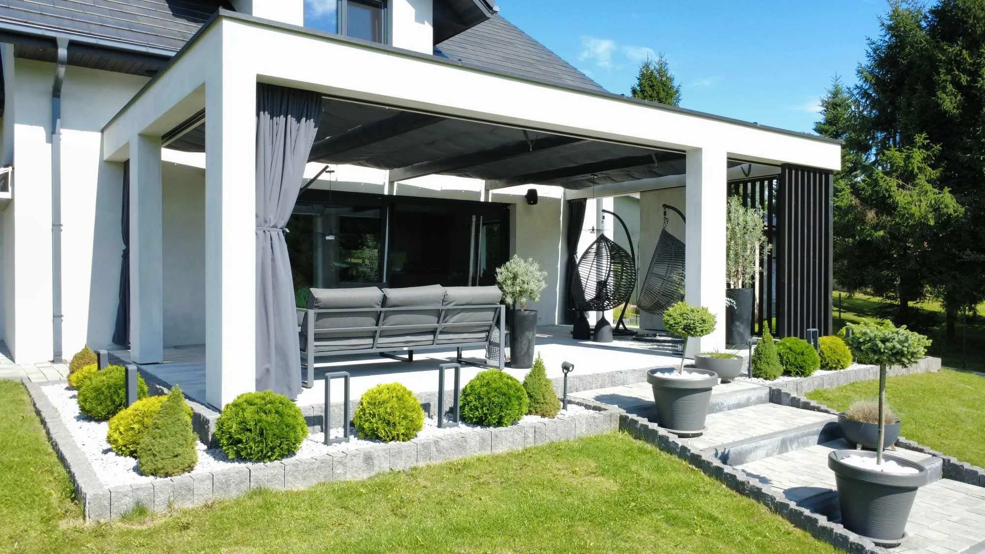 Modern backyard patio with outdoor furniture, hanging chairs, potted plants, surrounded by a neatly landscaped garden with green shrubs and trees.
