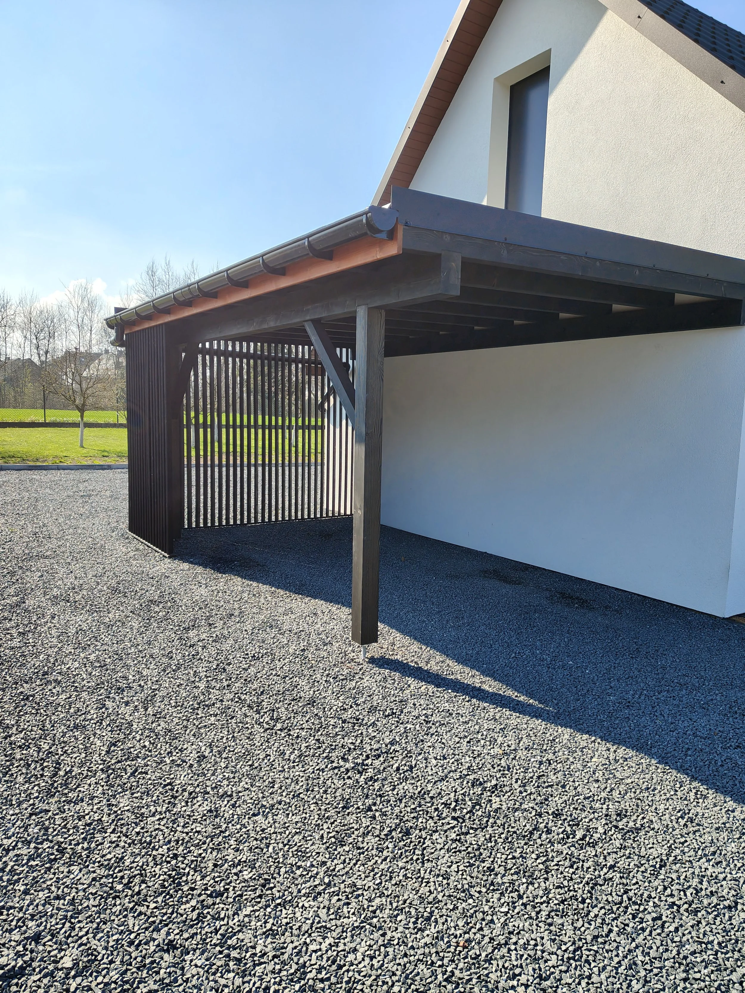 Exterior view of a modern house with a white wall, black window, and a wooden pergola structure on a gravel ground, with a grassy field and trees in the background under a clear blue sky.