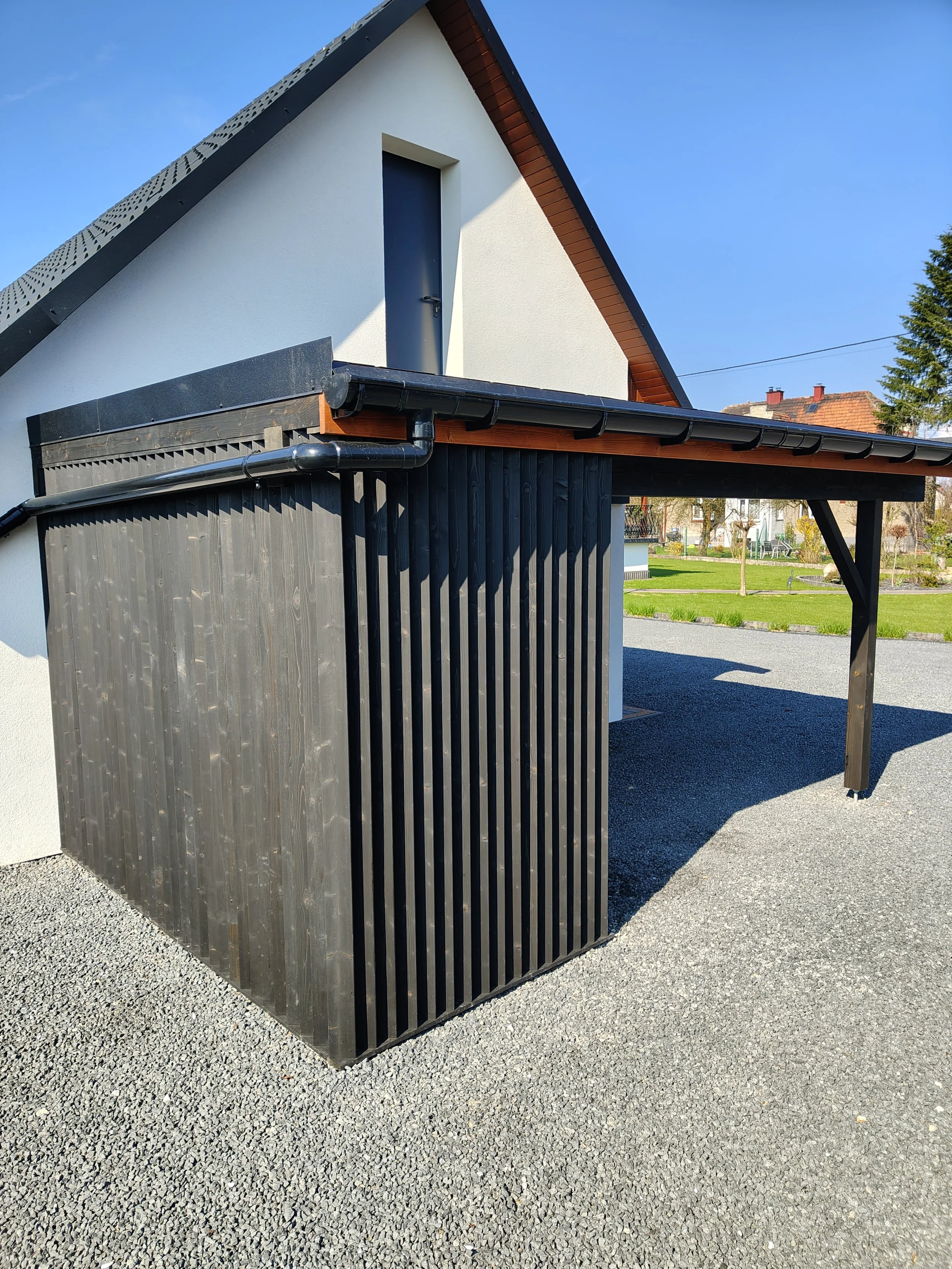 A modern house with a steep black roof, white walls, and a small window with a black frame. The house has a black gutter system and a wooden extension with vertical black wooden slats. The ground is covered with gray gravel, and there is a green lawn
